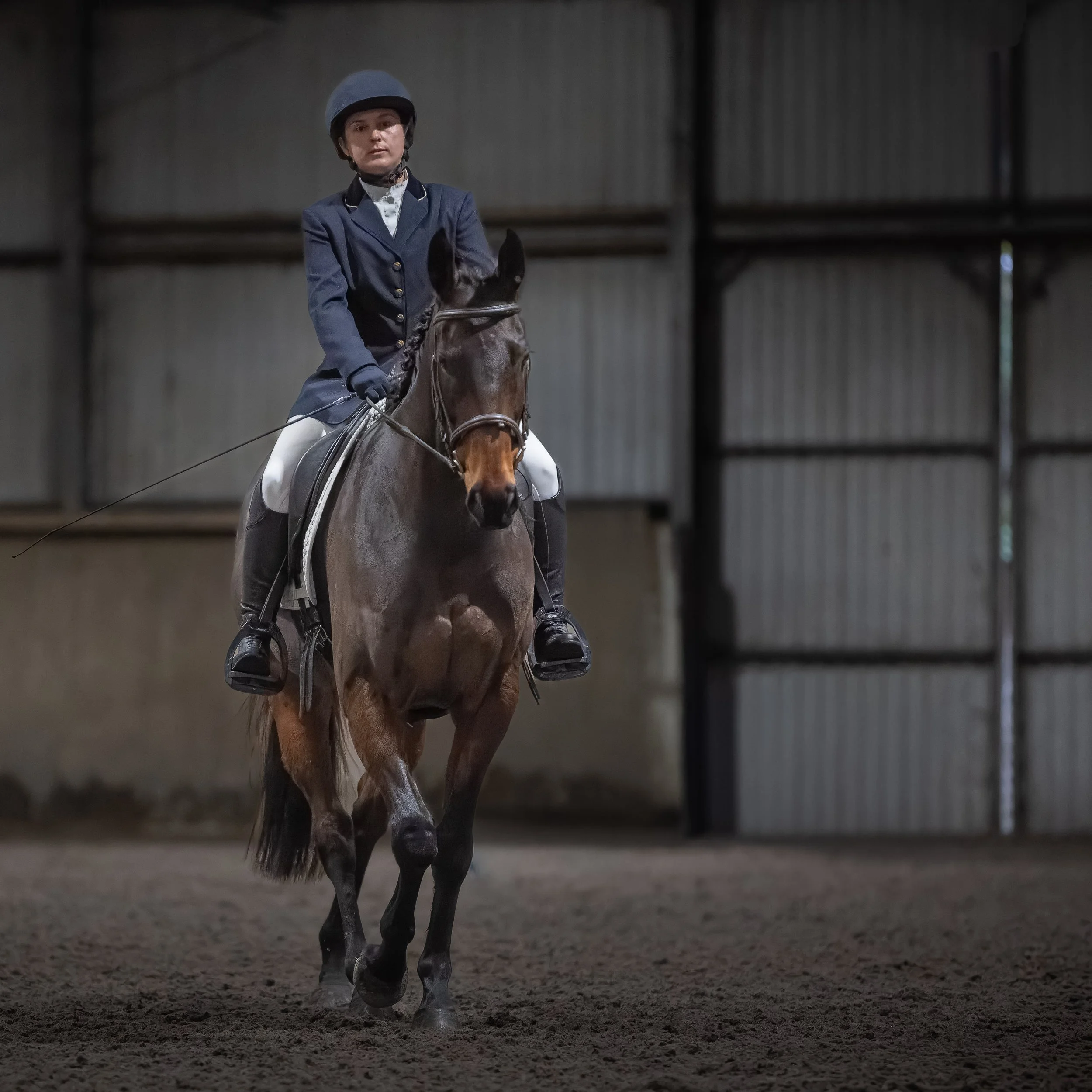 A woman wearing equestrian attire riding a brown horse inside an indoor riding arena.