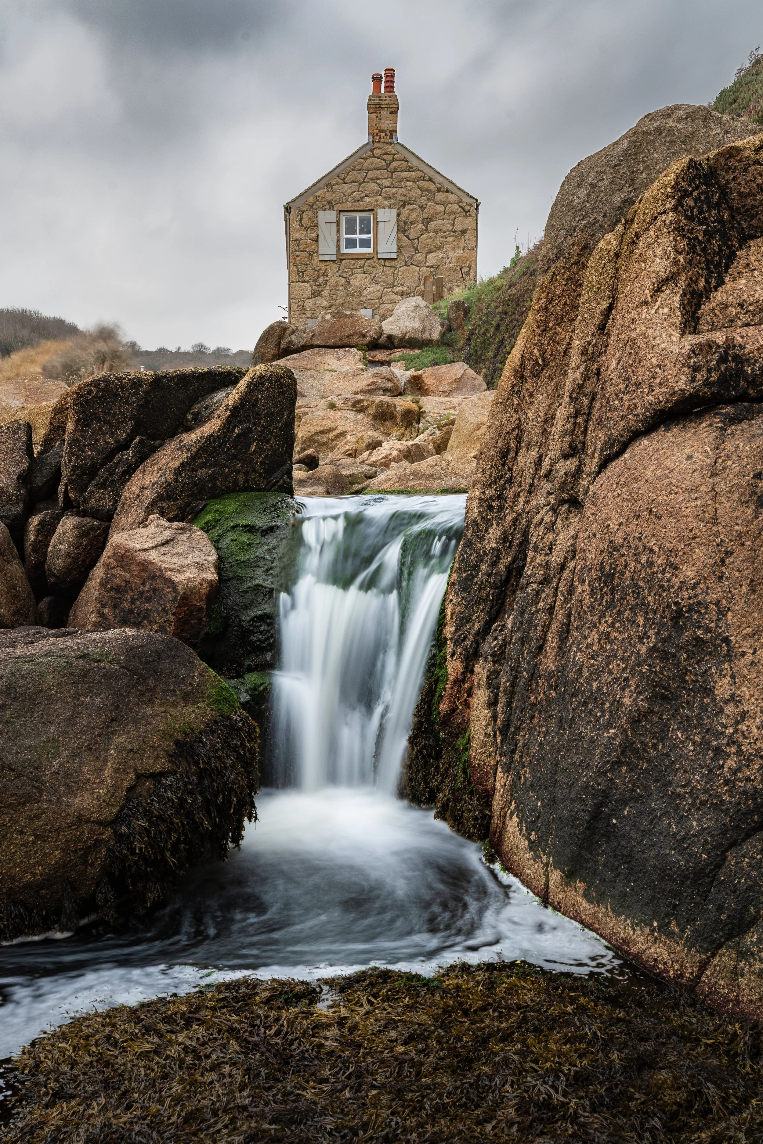 Stone house on a hill with water flowing over rocks in the foreground under cloudy sky.