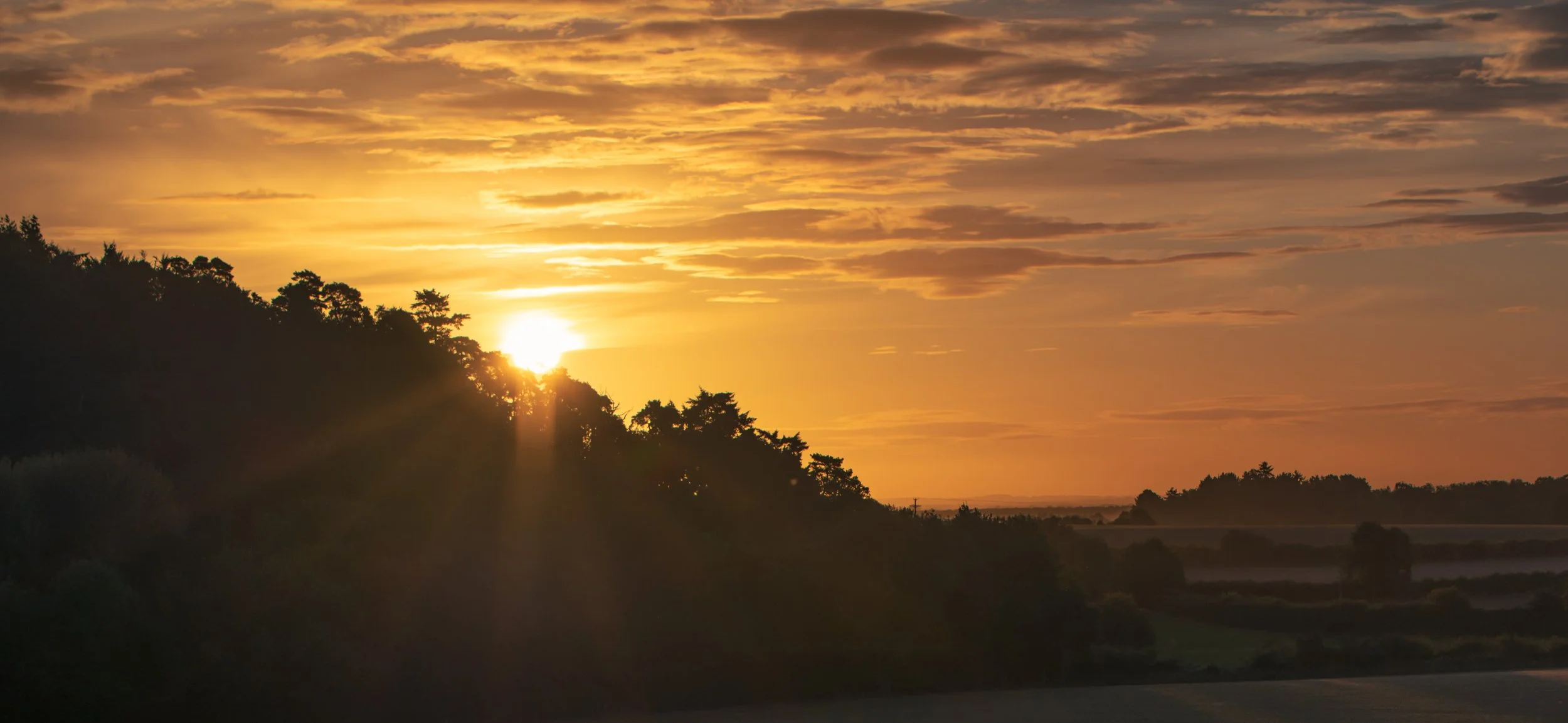 Sunset over a hilly landscape with trees and clouds in the sky.