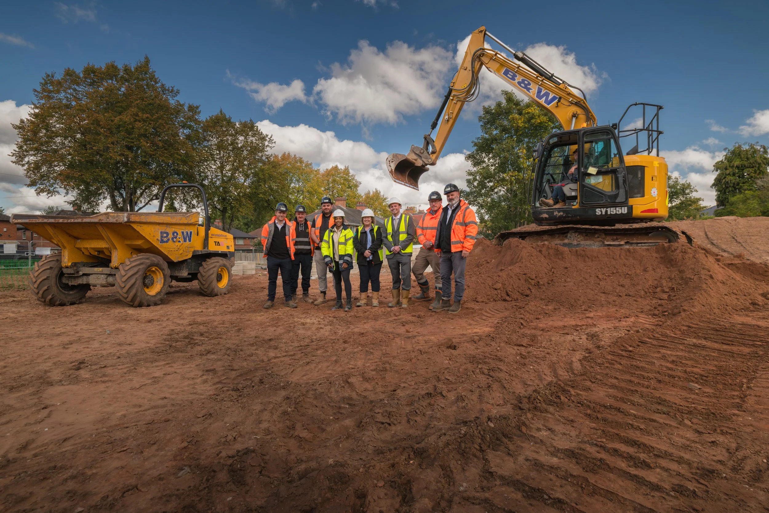 Construction workers wearing safety vests and helmets standing on a construction site with a small excavator and a dump truck under a partly cloudy sky.
