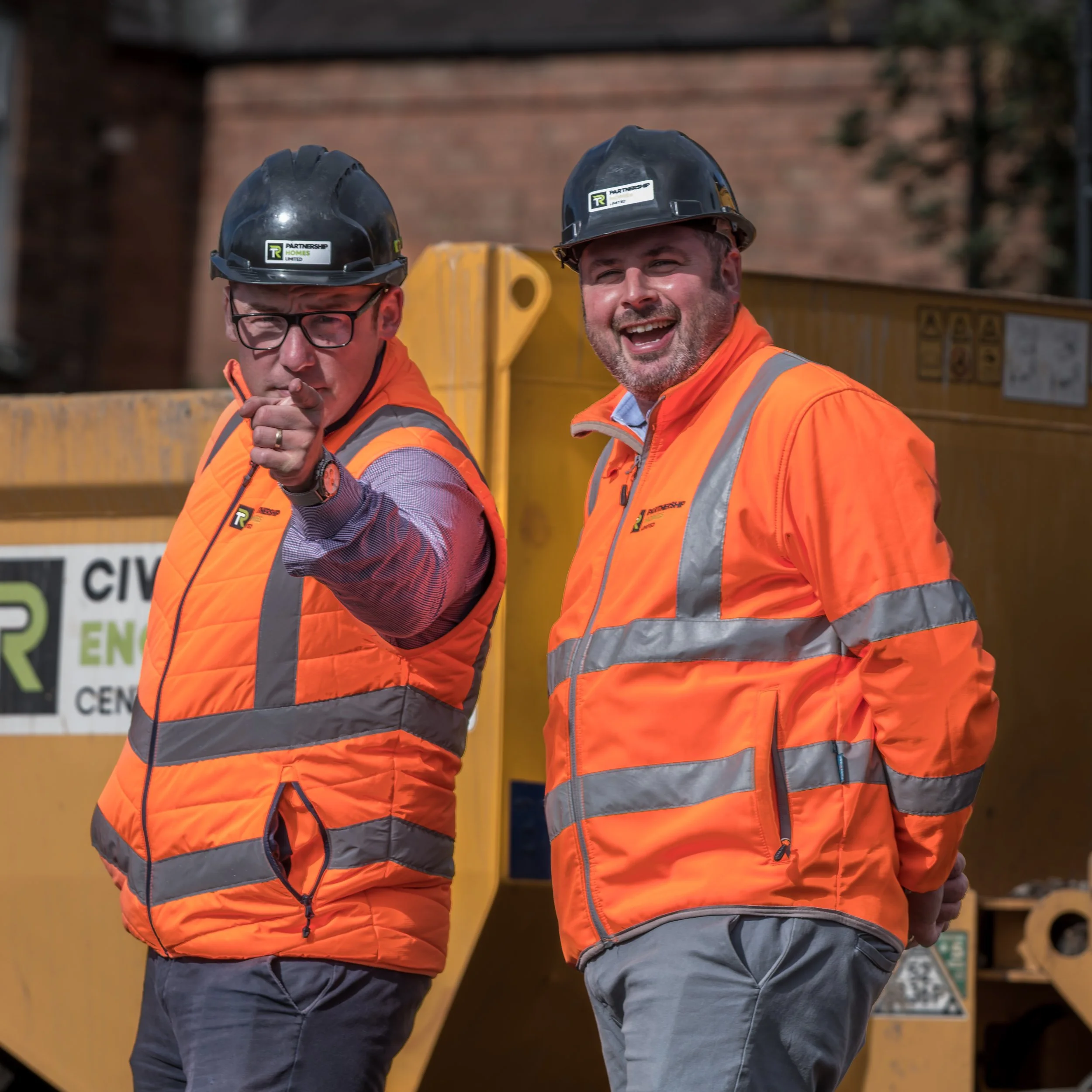 Two construction workers wearing orange safety vests and black hard hats, standing outdoors next to a yellow construction vehicle. One is pointing towards the camera, and both are smiling.