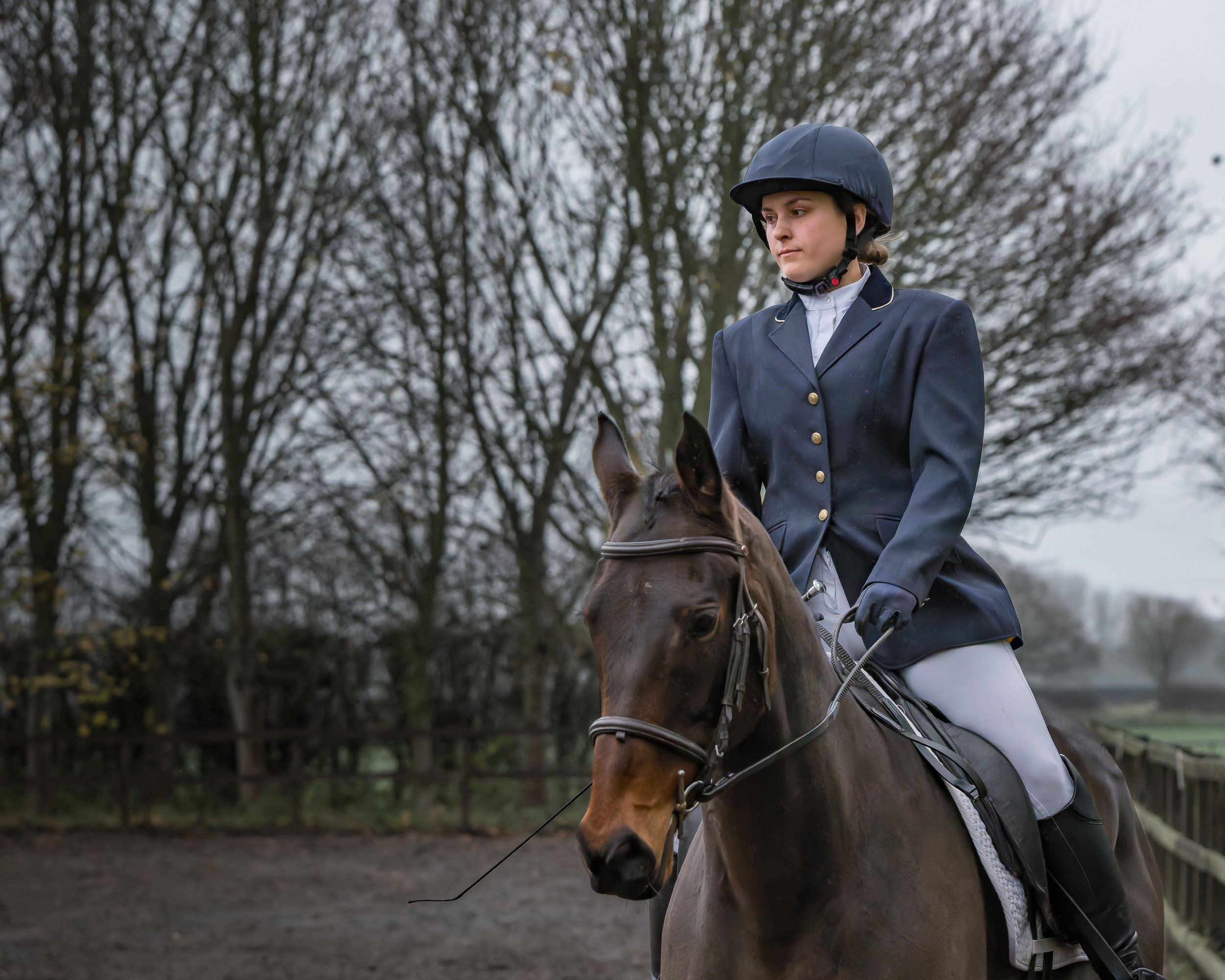 A young woman in formal equestrian attire, including a helmet, navy blazer, and riding gloves, riding a brown horse in an outdoor riding arena with leafless trees and a wooden fence in the background.