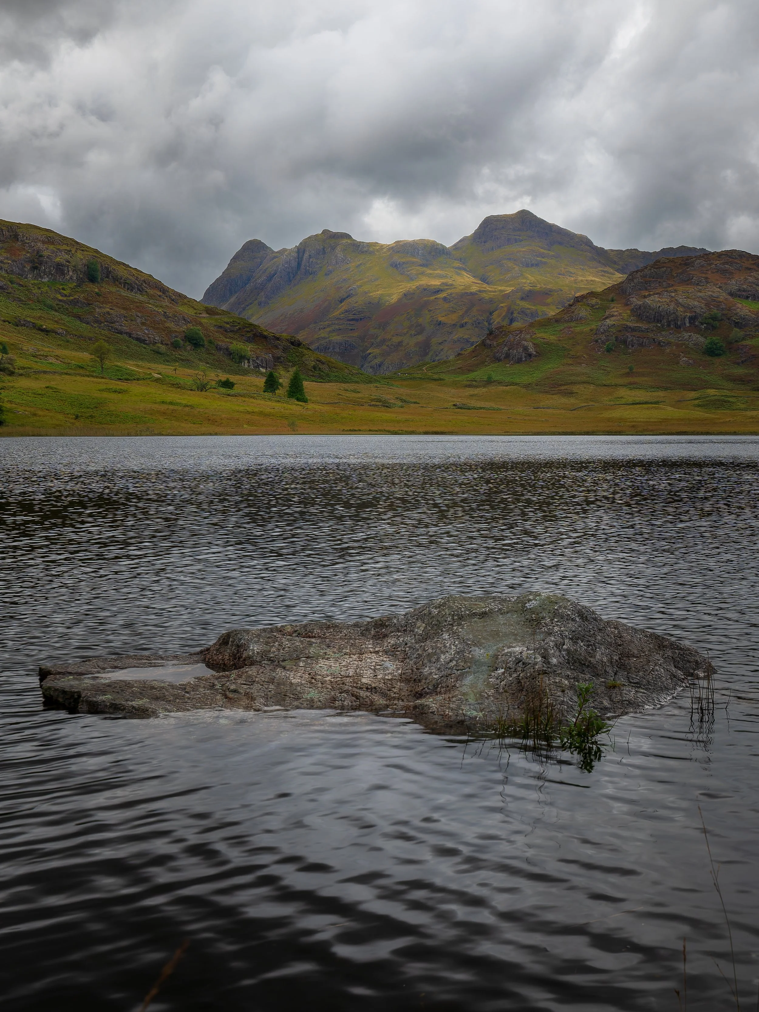 A lake with a partially submerged large rock in the foreground, surrounded by calm water, with green hills and mountains in the background under a cloudy sky.