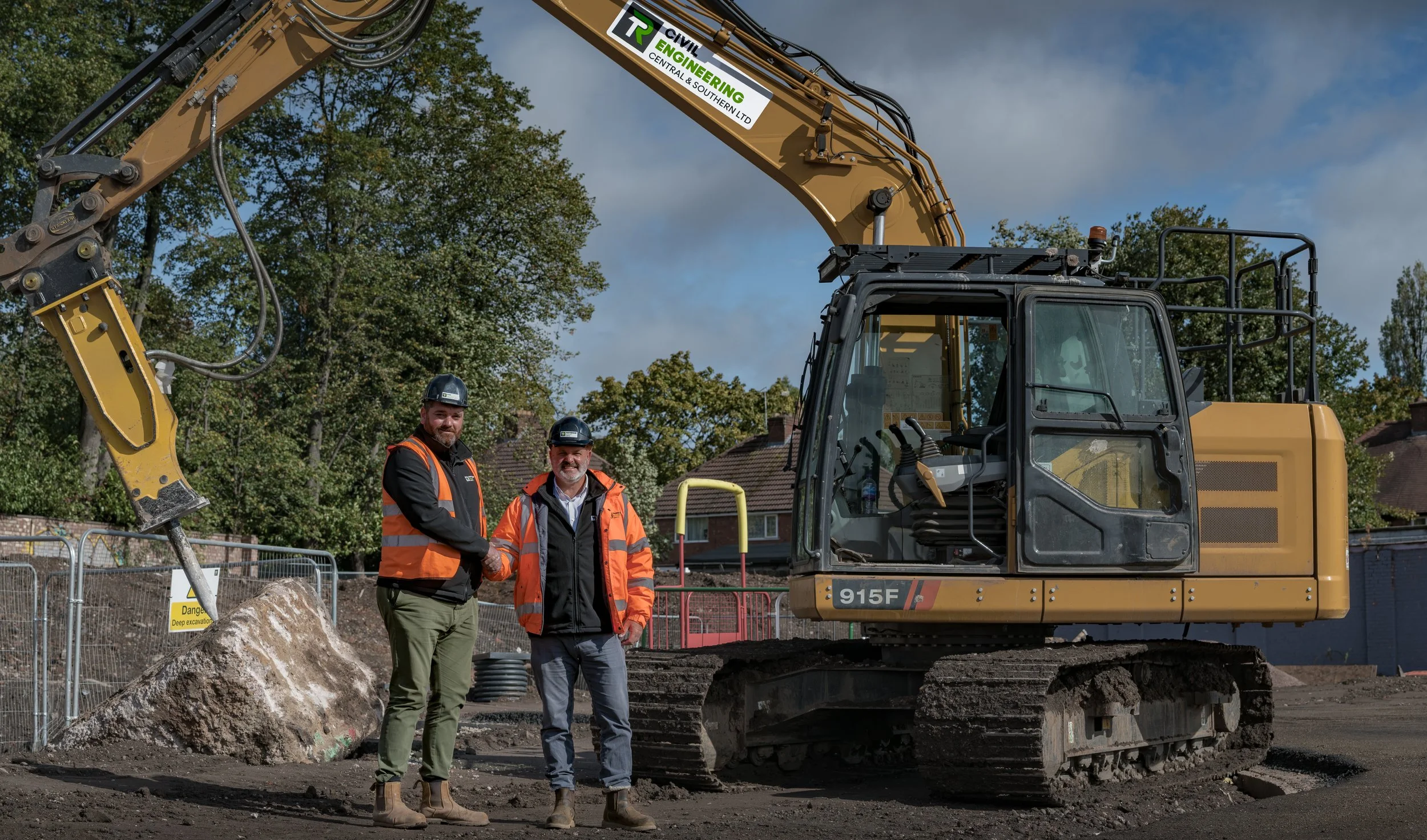 Two construction workers shaking hands in front of a large excavator at a construction site, with trees and houses in the background.