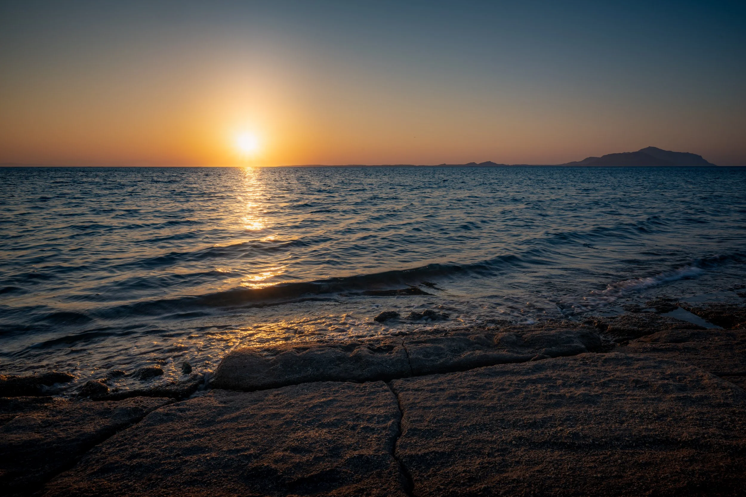 Sunset over the ocean with rocky shoreline in the foreground and an island in the distance.