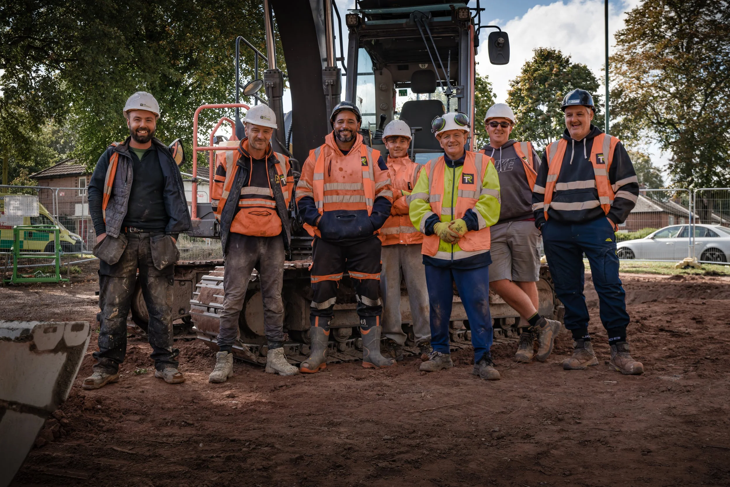 Group of construction workers standing in front of an excavator on a construction site, wearing safety gear including helmets, vests, and boots.