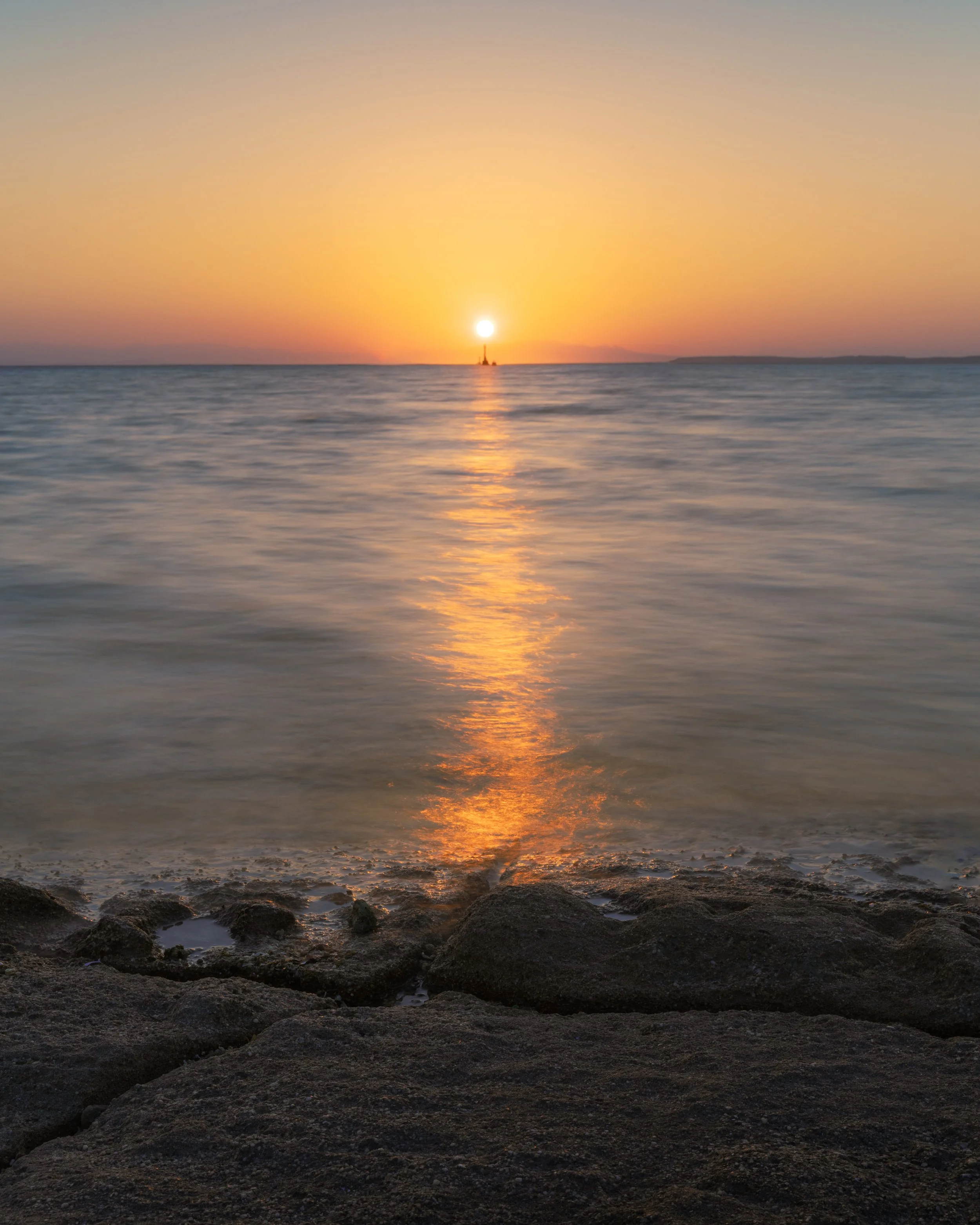 Sunset over calm ocean with a reflection on the water and rocks in the foreground.