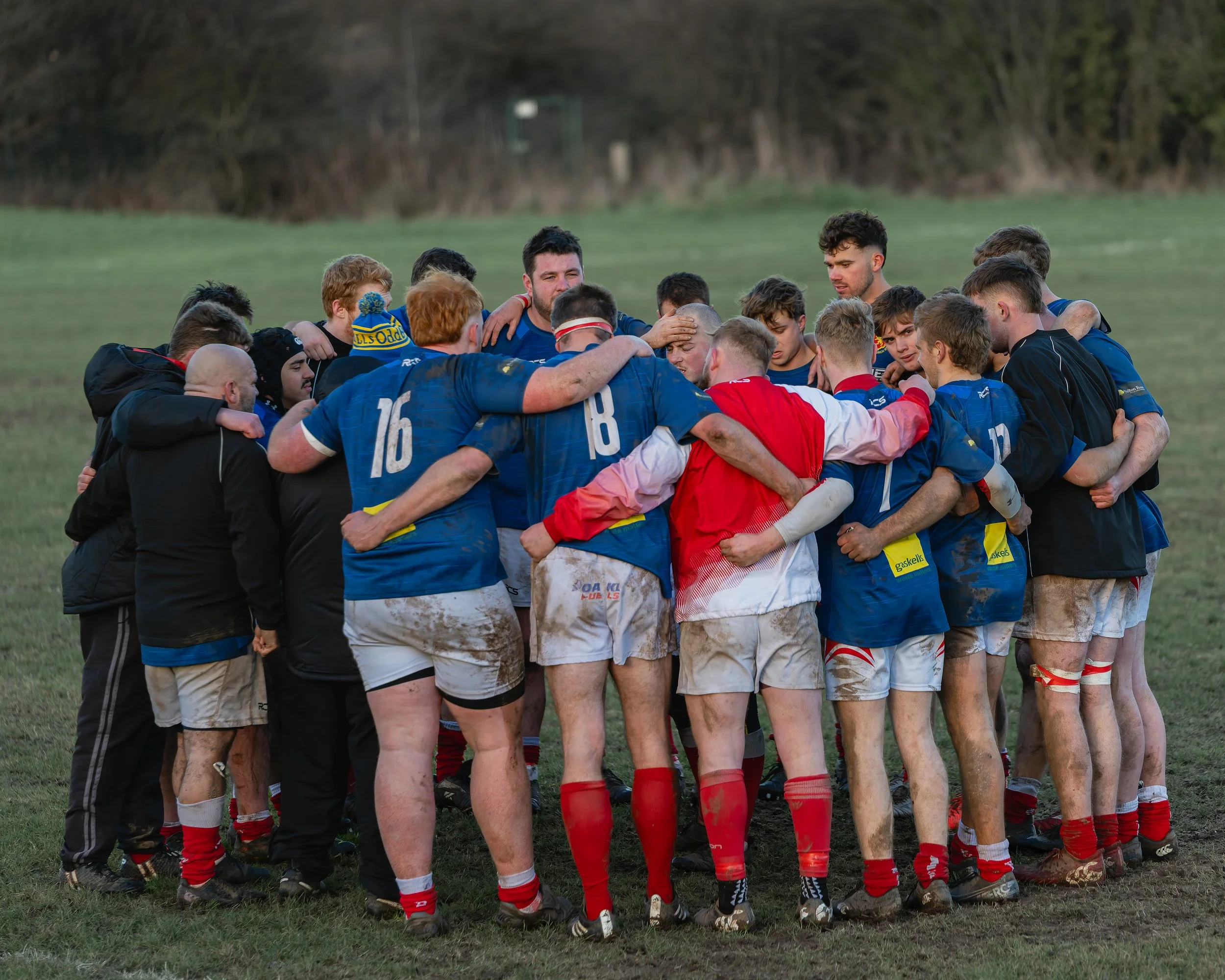 A rugby team huddles together on a grassy field, showing team unity and focus, with some players covered in dirt, indicating a recent match or practice.
