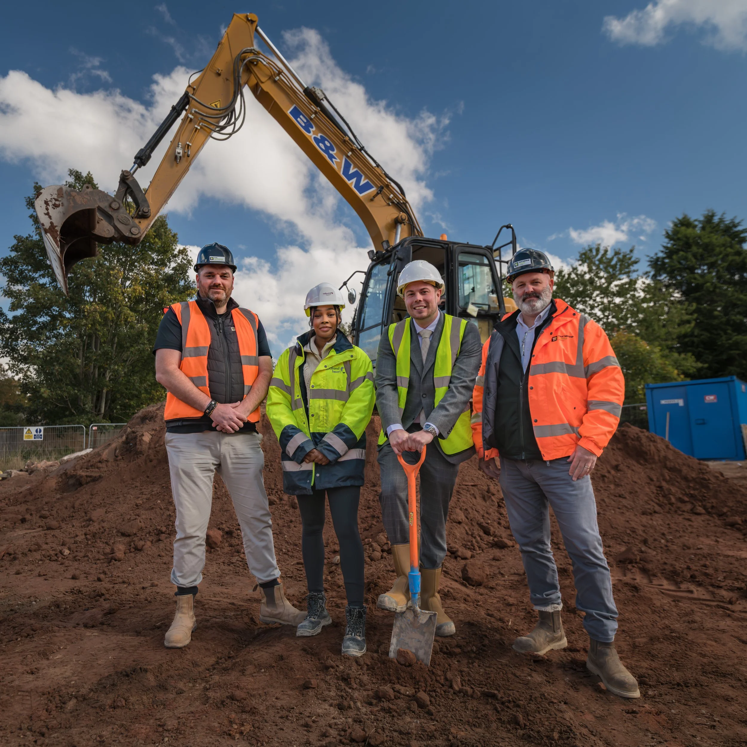 Group of four construction professionals standing on a construction site with dirt, wearing safety gear including helmets and reflective vests, in front of a large excavator.