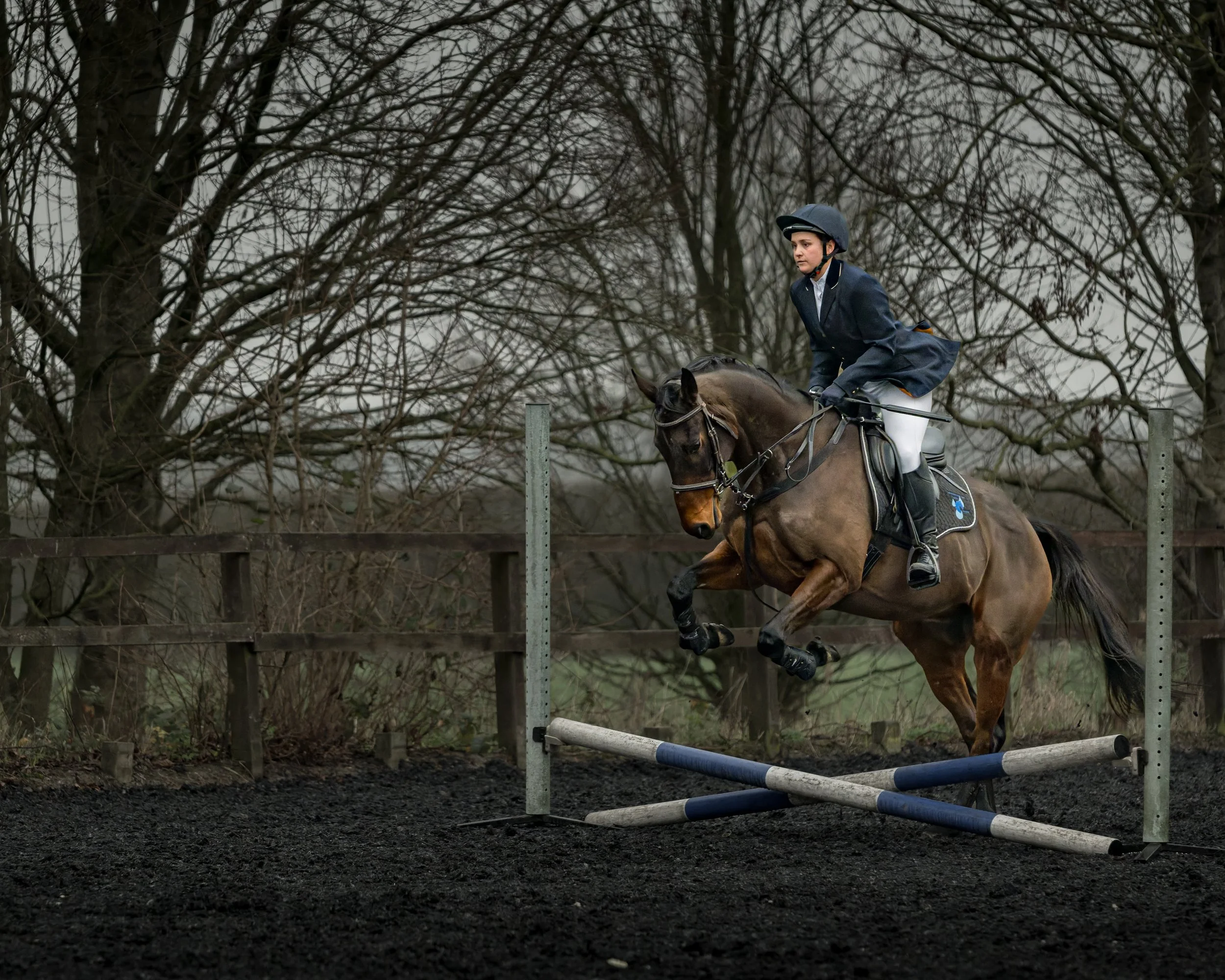 A person wearing riding gear and a helmet riding a horse over a jump in an outdoor equestrian arena.