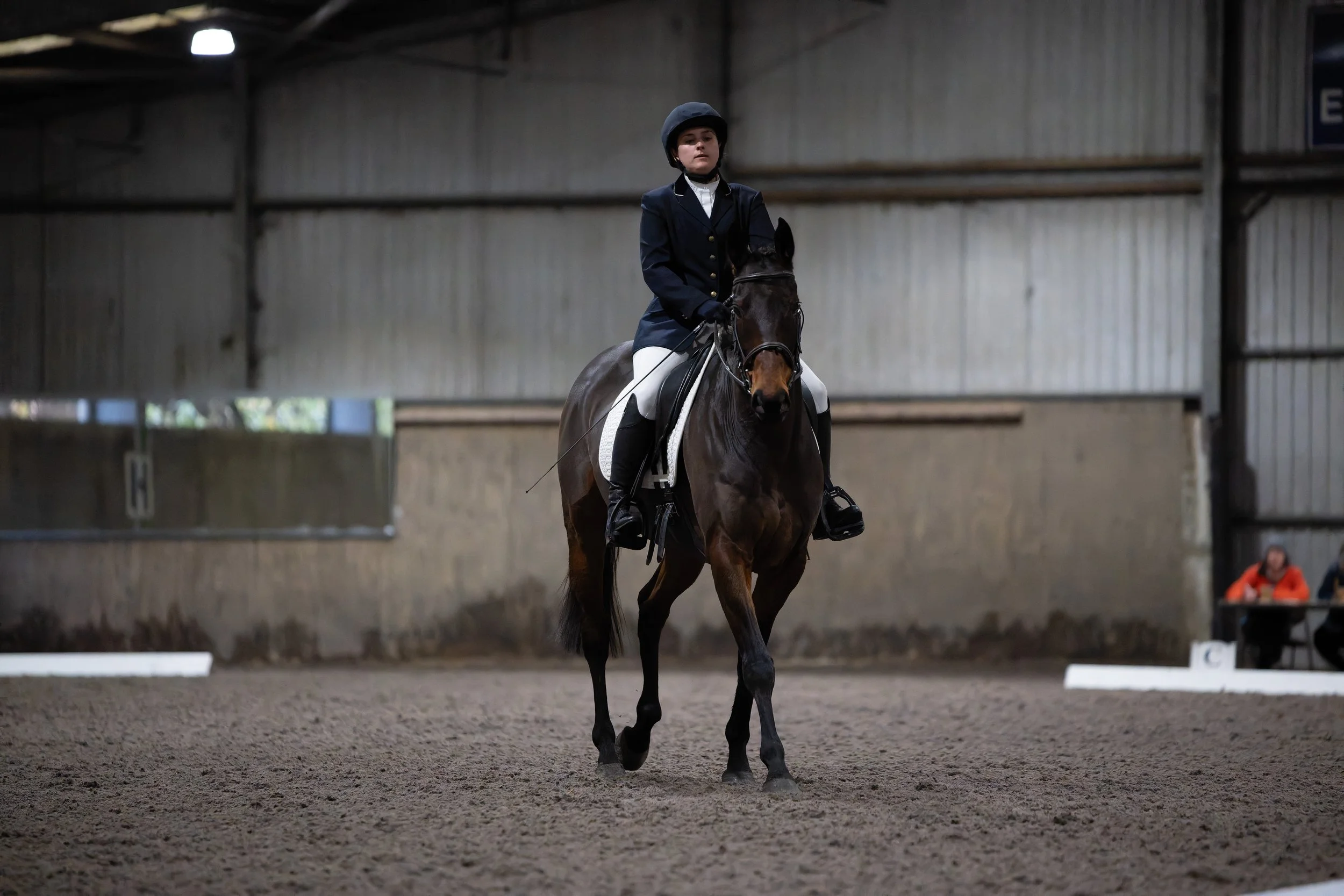 A young female equestrian dressed in formal riding attire, including a helmet, riding a dark horse inside an indoor arena.