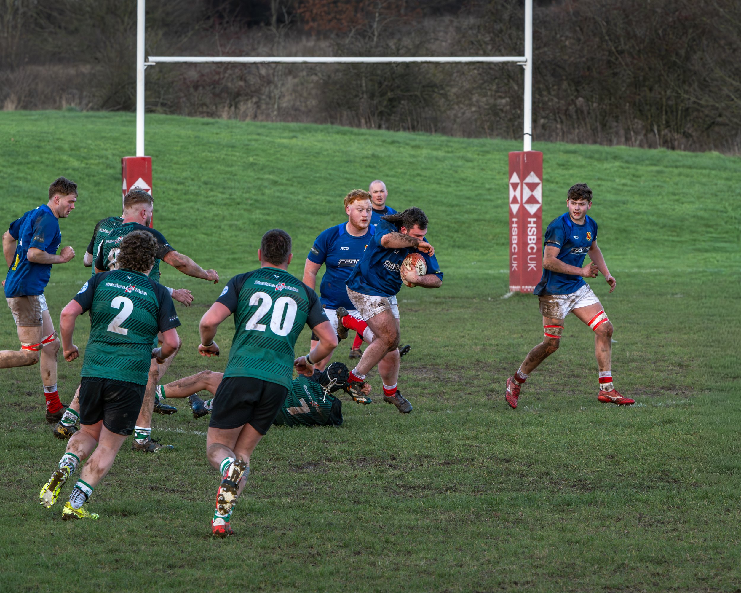 A rugby game with players running and tackling on a muddy field, with goalposts and a grassy hill in the background.