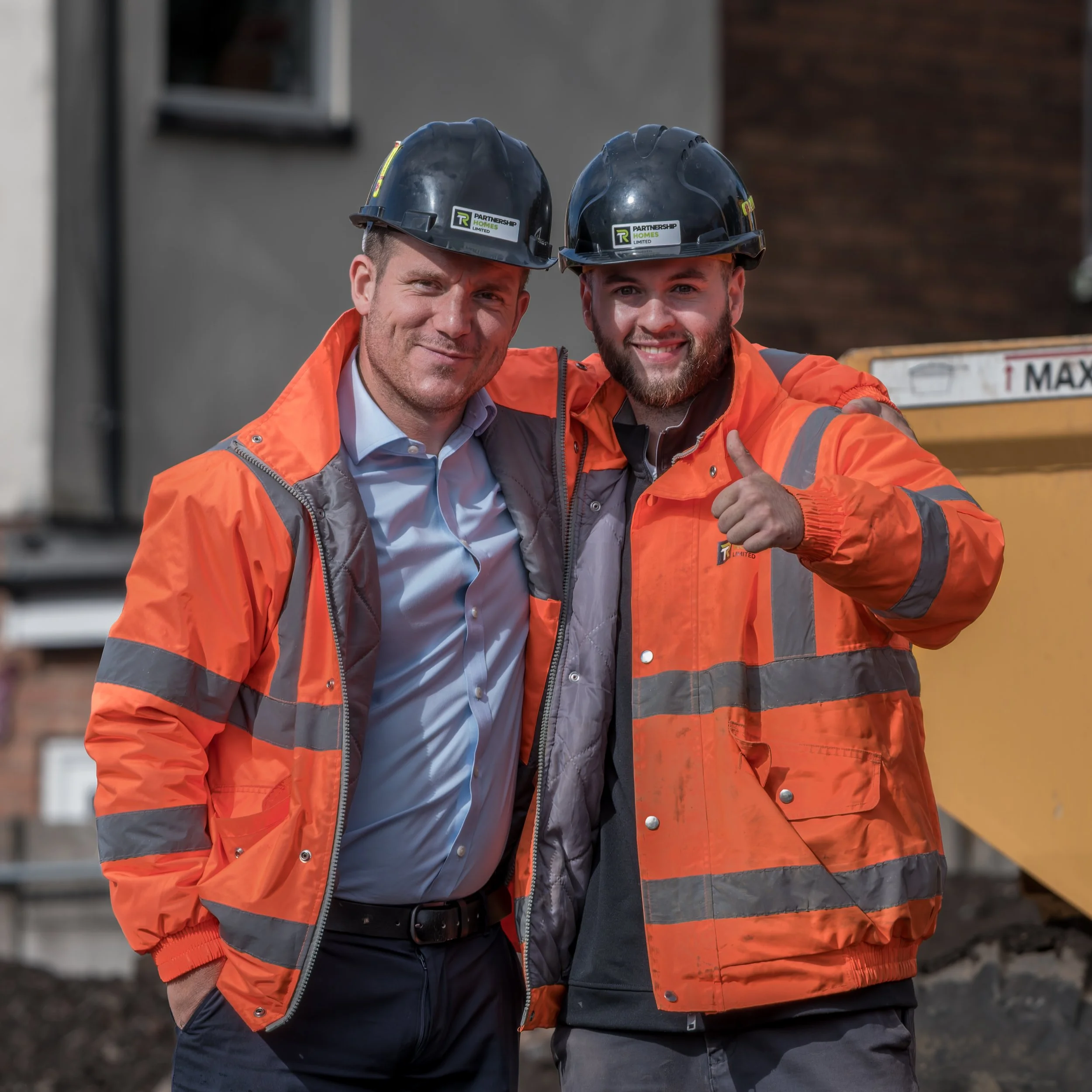 Two construction workers in orange safety jackets and black helmets smiling and posing for the camera, one giving a thumbs up, at a construction site.