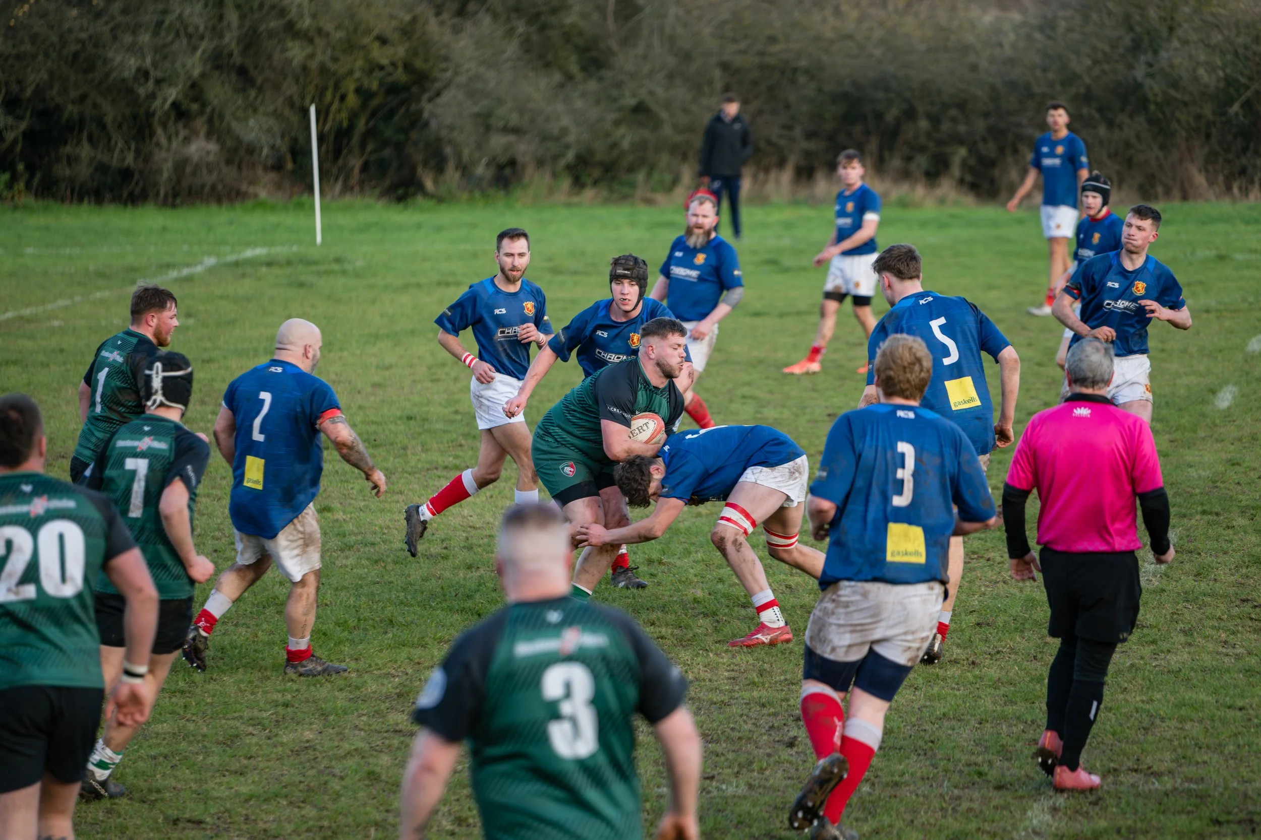 Rugby players in blue and green jerseys competing for the ball on a grassy field, with referees and spectators observing.