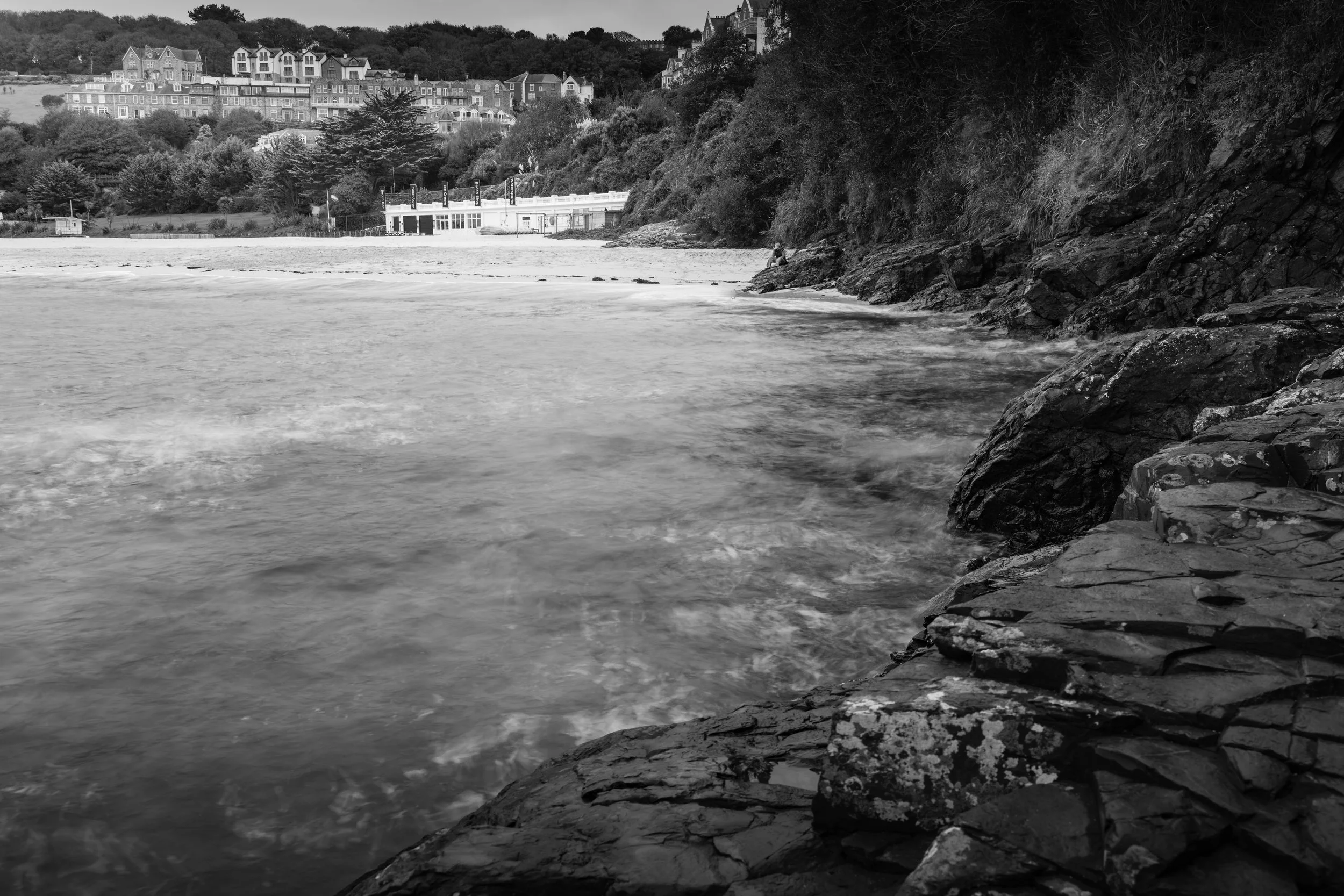 A black and white photo of a rocky coastal area with water in the foreground, a sandy beach and buildings on the far side, and a hillside with houses and trees in the background.
