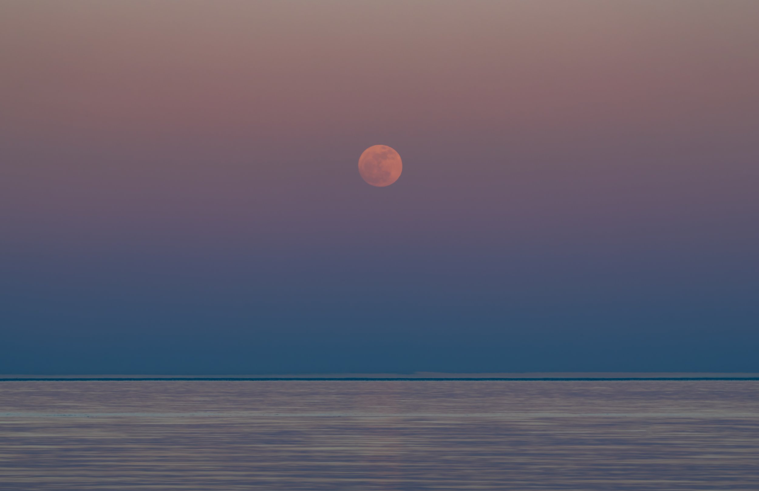 Full moon over a calm ocean at sunset with a pink and purple sky.