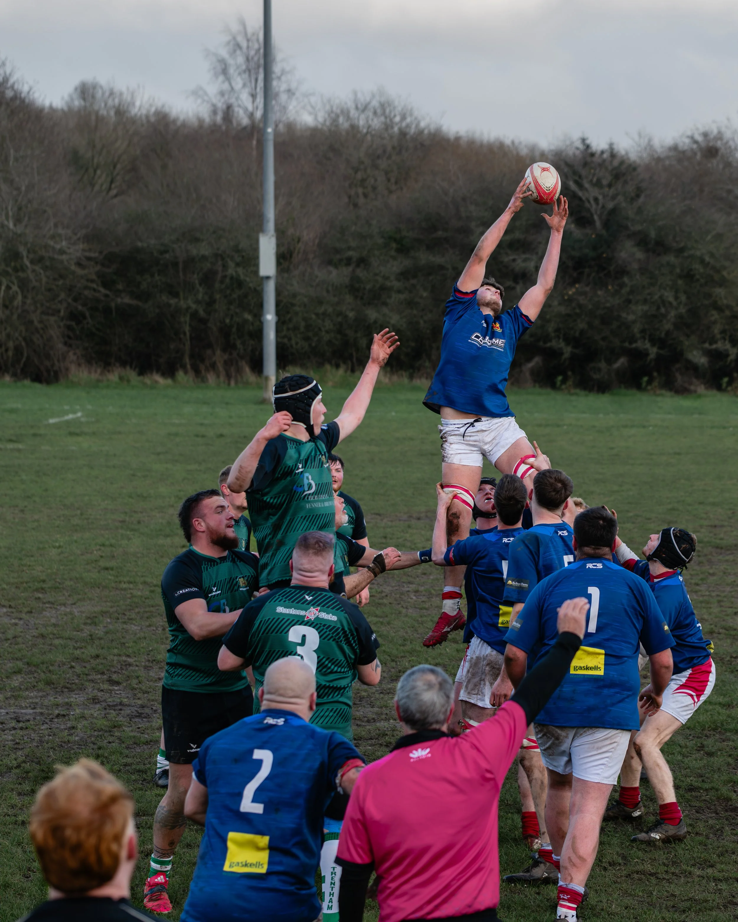 Rugby players competing in a lineout on a grassy field, with one player in blue jumping to catch the ball, others in green and blue uniform supporting, and a referee in pink observing.