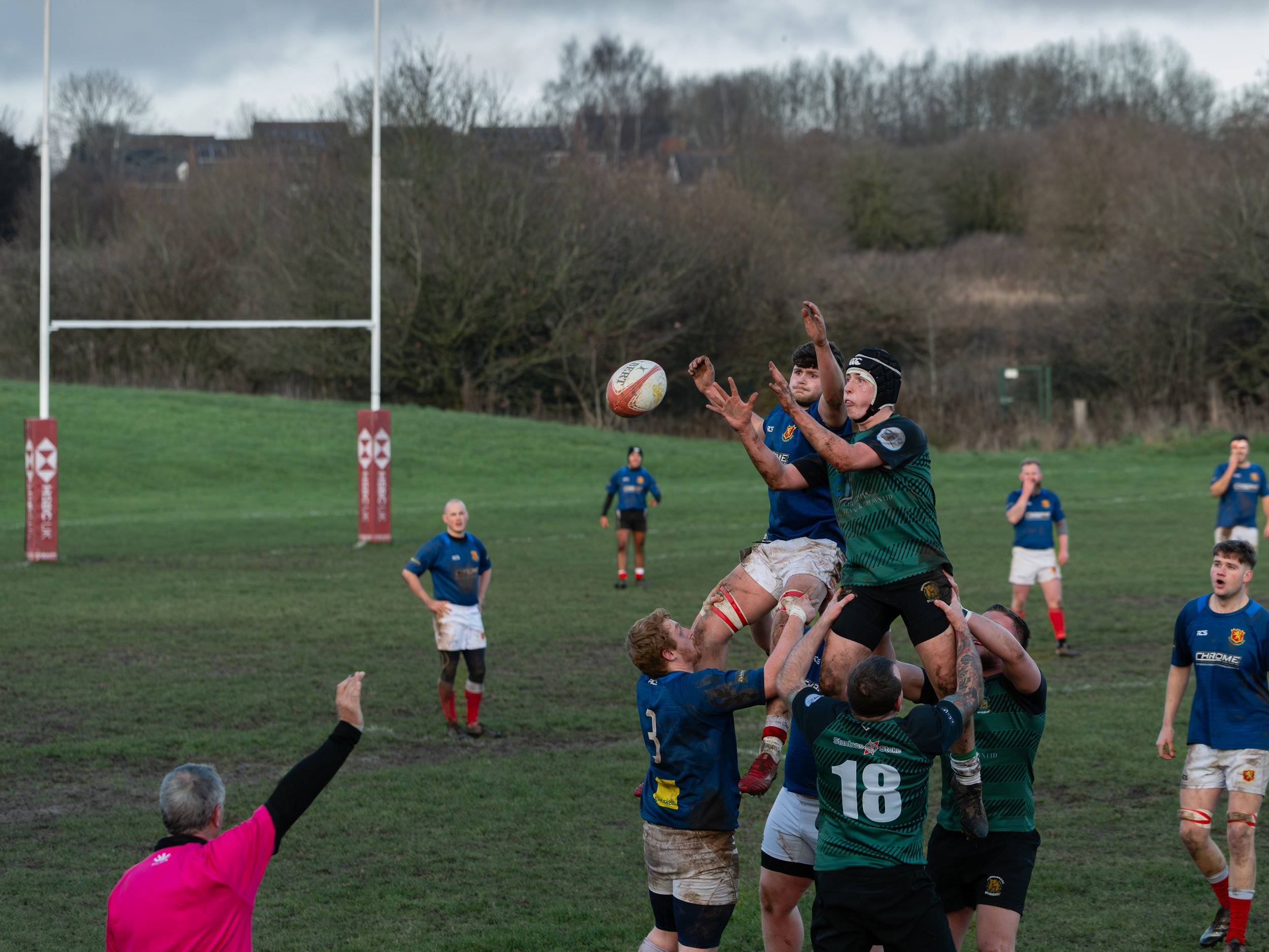 Rugby players in a lineout competing to catch the ball during a match, with players from both teams jumping for the ball on a grassy field, and a referee in a pink jersey signaling.