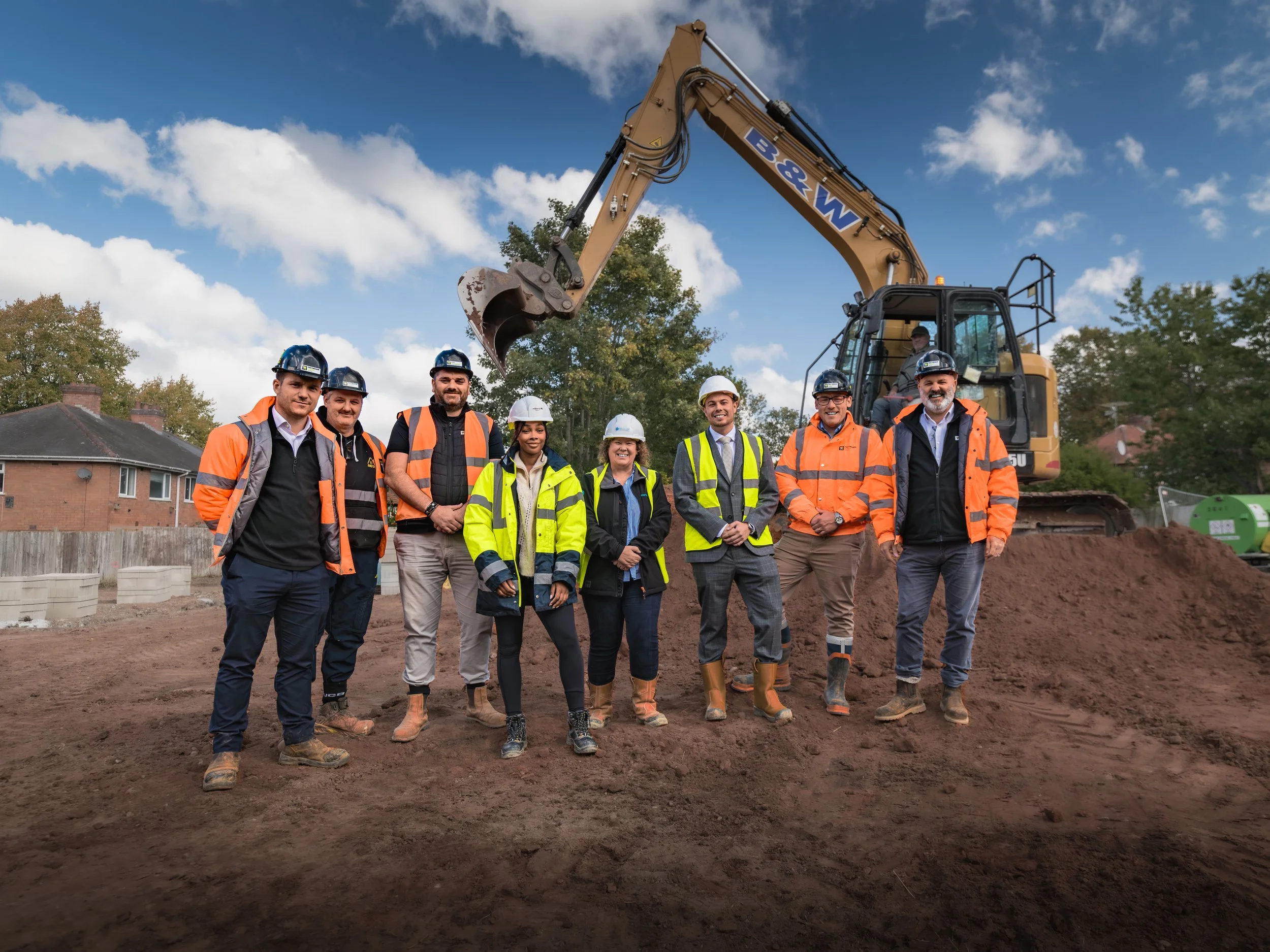 A group of nine construction workers and engineers standing on a dirt construction site, posing for a photo with an excavator behind them and a partly cloudy sky overhead.