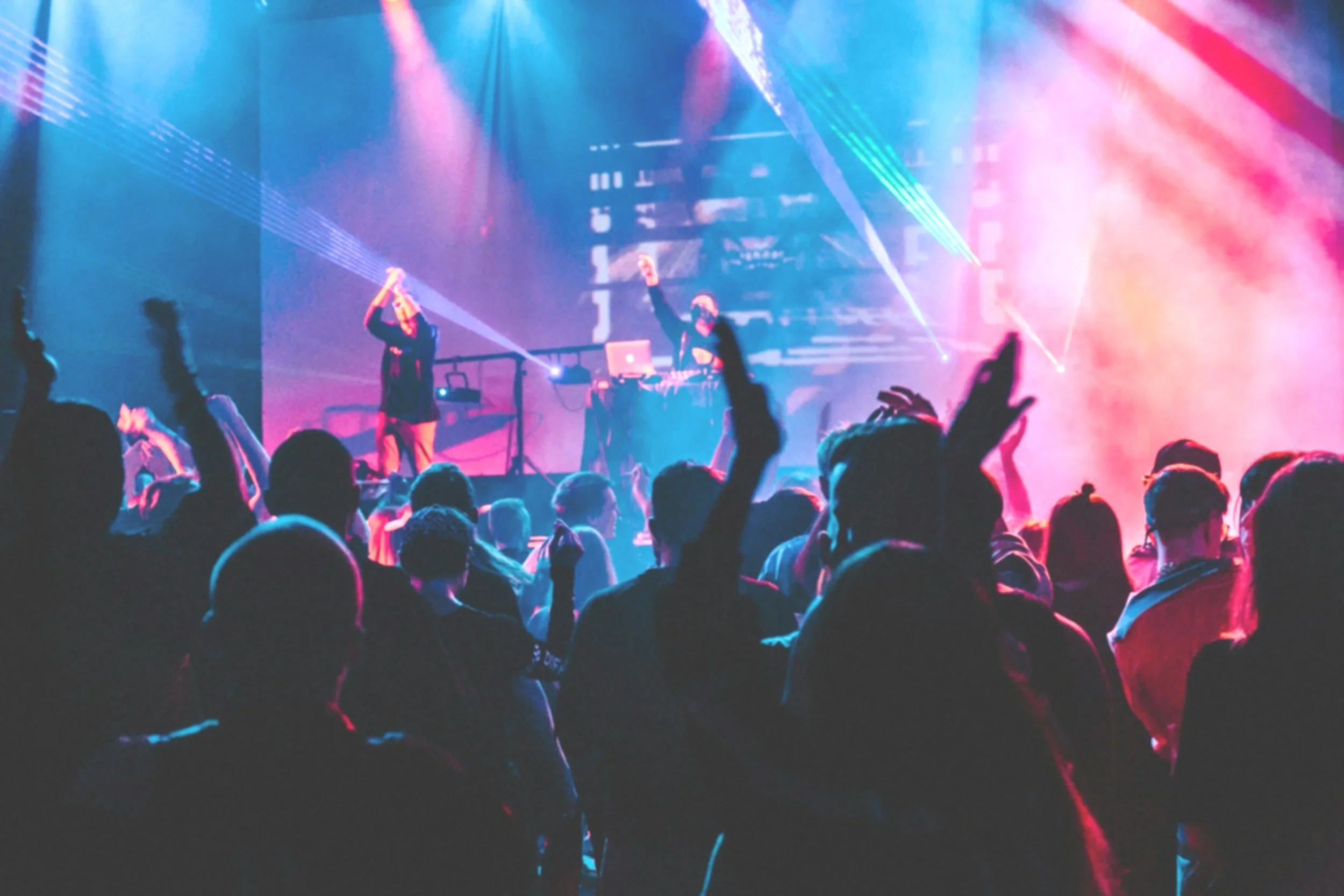 Crowd at a concert or music event with colorful lights, DJ and performers on stage, people dancing and raising hands.