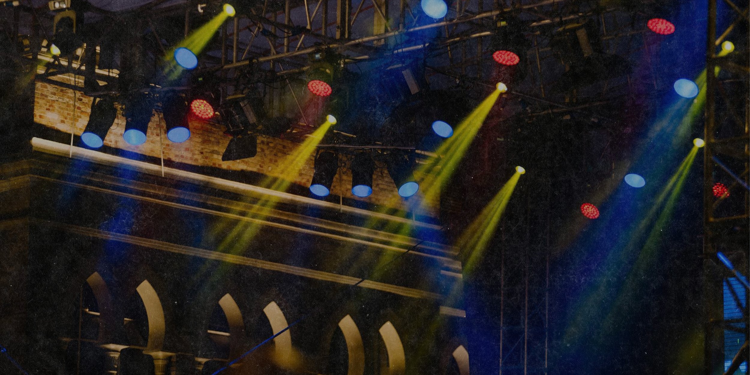 Stage with colorful lighting, including yellow, blue, and red spotlights, featuring a brick wall and scaffolding structures.