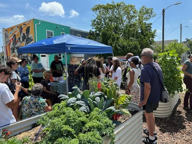 People gathered at an outdoor farmers market with fresh vegetables, under a blue canopy, in front of colorful murals and green trees.