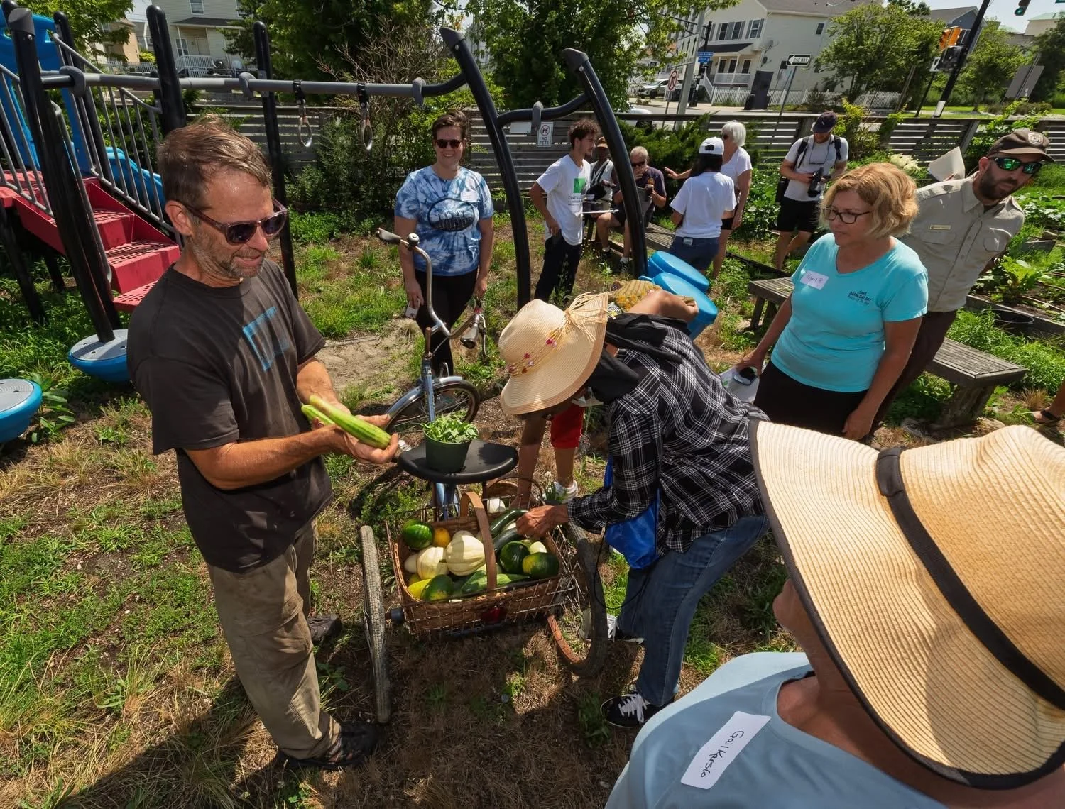 People gathered in a community garden during daytime, with a man holding zucchini and others looking at produce in a basket, surrounded by green plants, benches, and playground equipment.