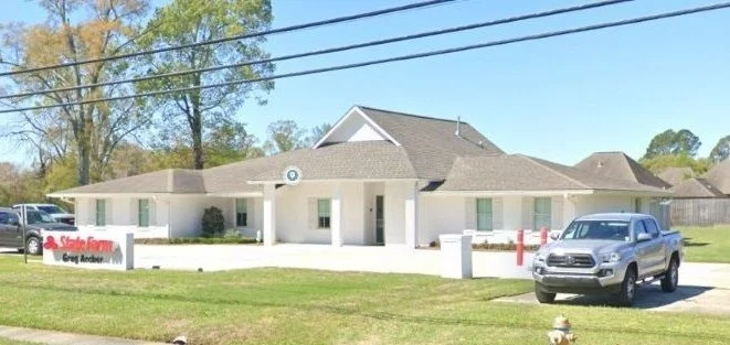 Single-story commercial building with a gray roof, surrounded by green lawn, parking lot with cars, and a sign that reads 'State Farm' with additional text that is not fully visible.
