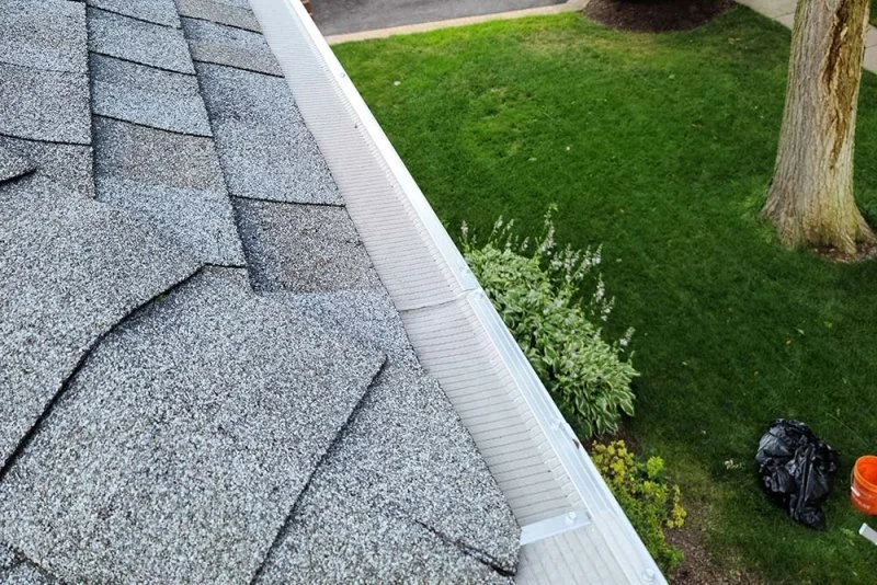 View from a roof showing gray shingles, roof gutter, and a yard with green grass and a tree.