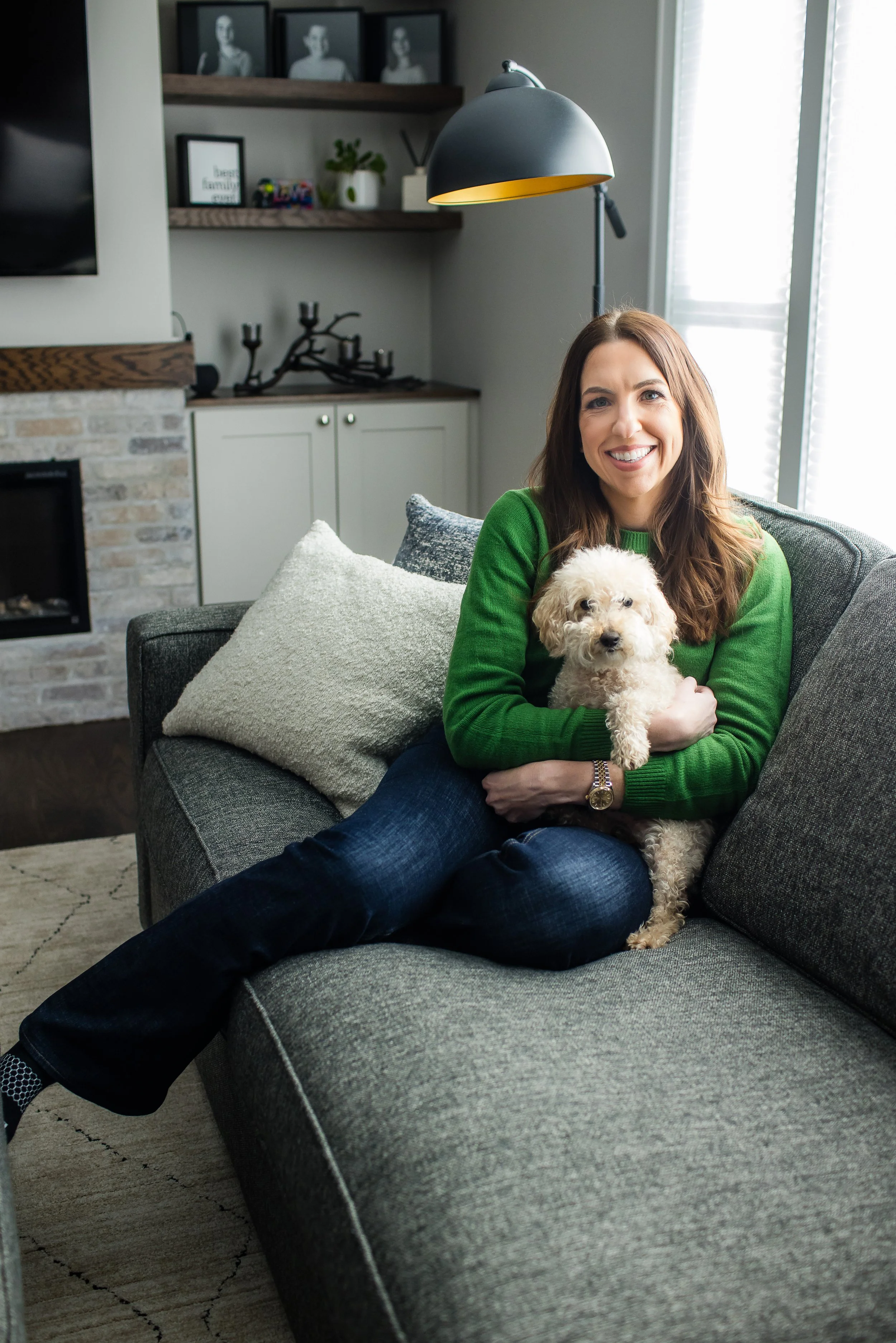 A smiling woman with brown hair in a green sweater sitting on a gray sofa holding a small curly-haired white dog in a modern living room with a brick fireplace, decorative pillows, and windows with blinds.