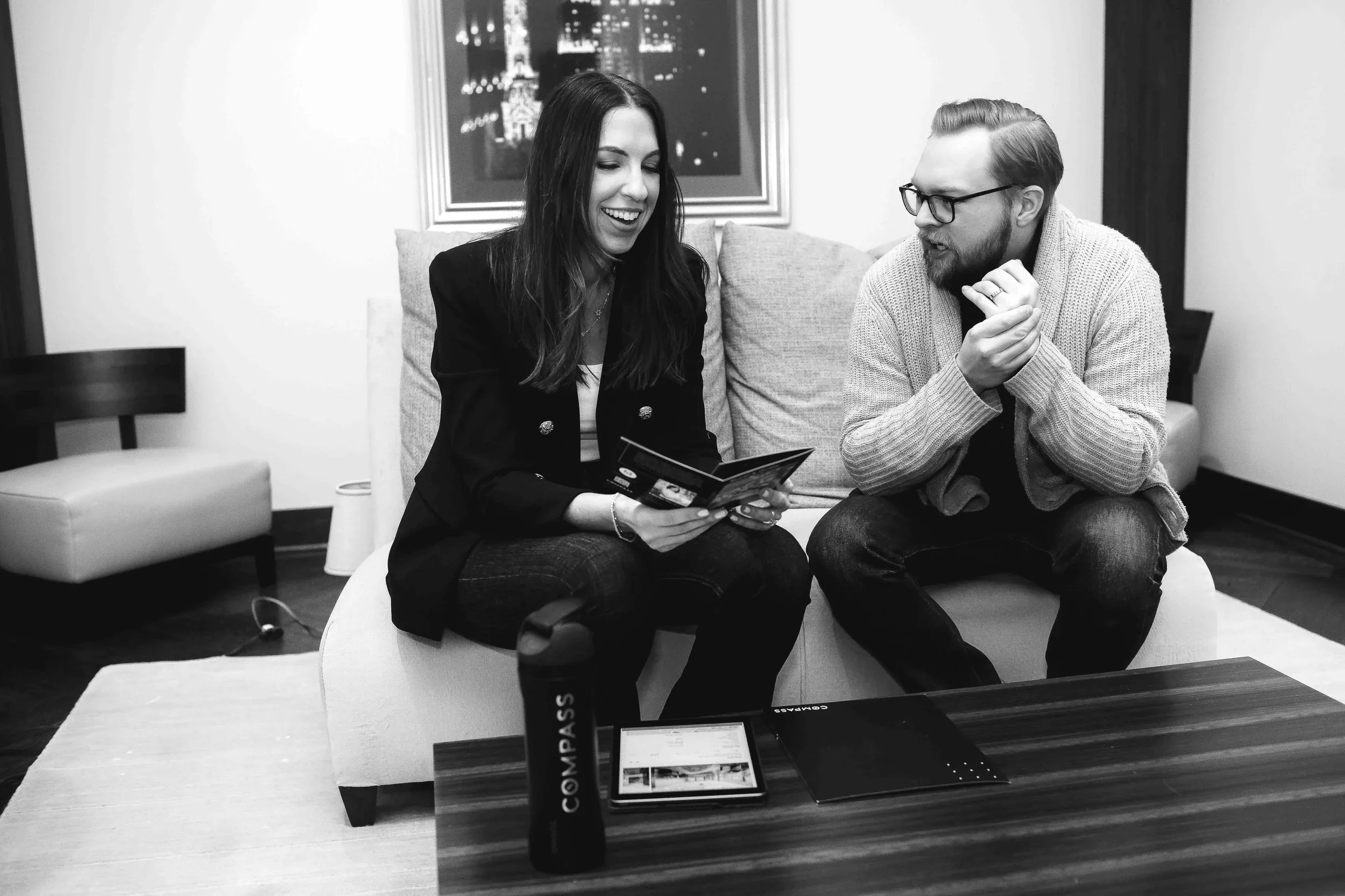 A woman and a man sitting on a couch in a living room, smiling and looking at a pamphlet or magazine together, with a framed picture behind them and a table in front of them.