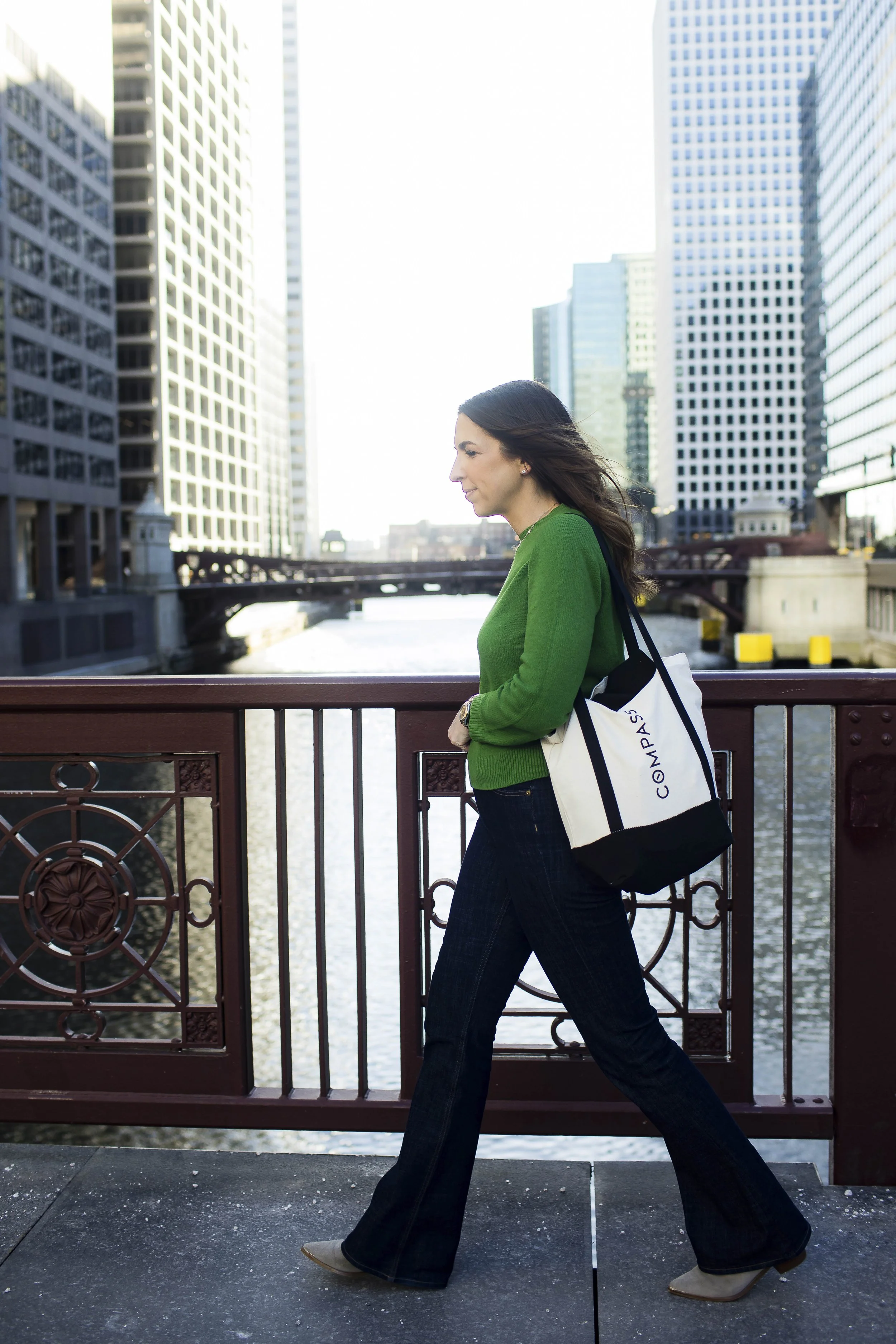 A woman walking on a city bridge near water, wearing a green sweater, dark jeans, and beige shoes, carrying a white and black bag labeled 'COMPASS' with tall office buildings in the background.