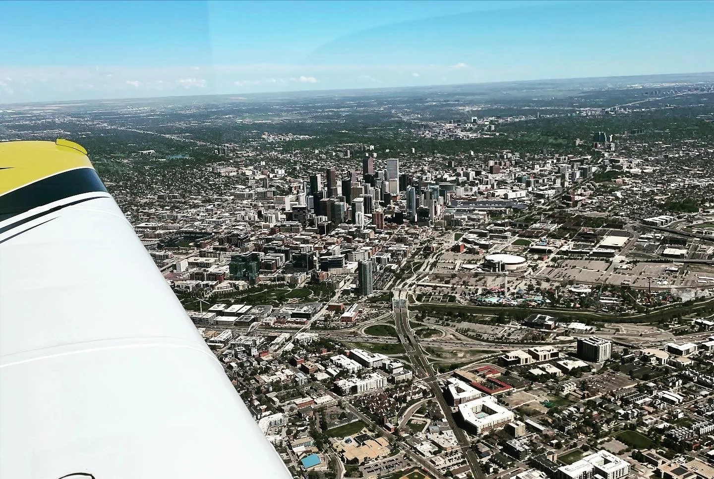 Aerial view of a city with a concentration of skyscrapers, winding roads, and various buildings, taken from a small aircraft in flight, with part of the aircraft visible on the left.