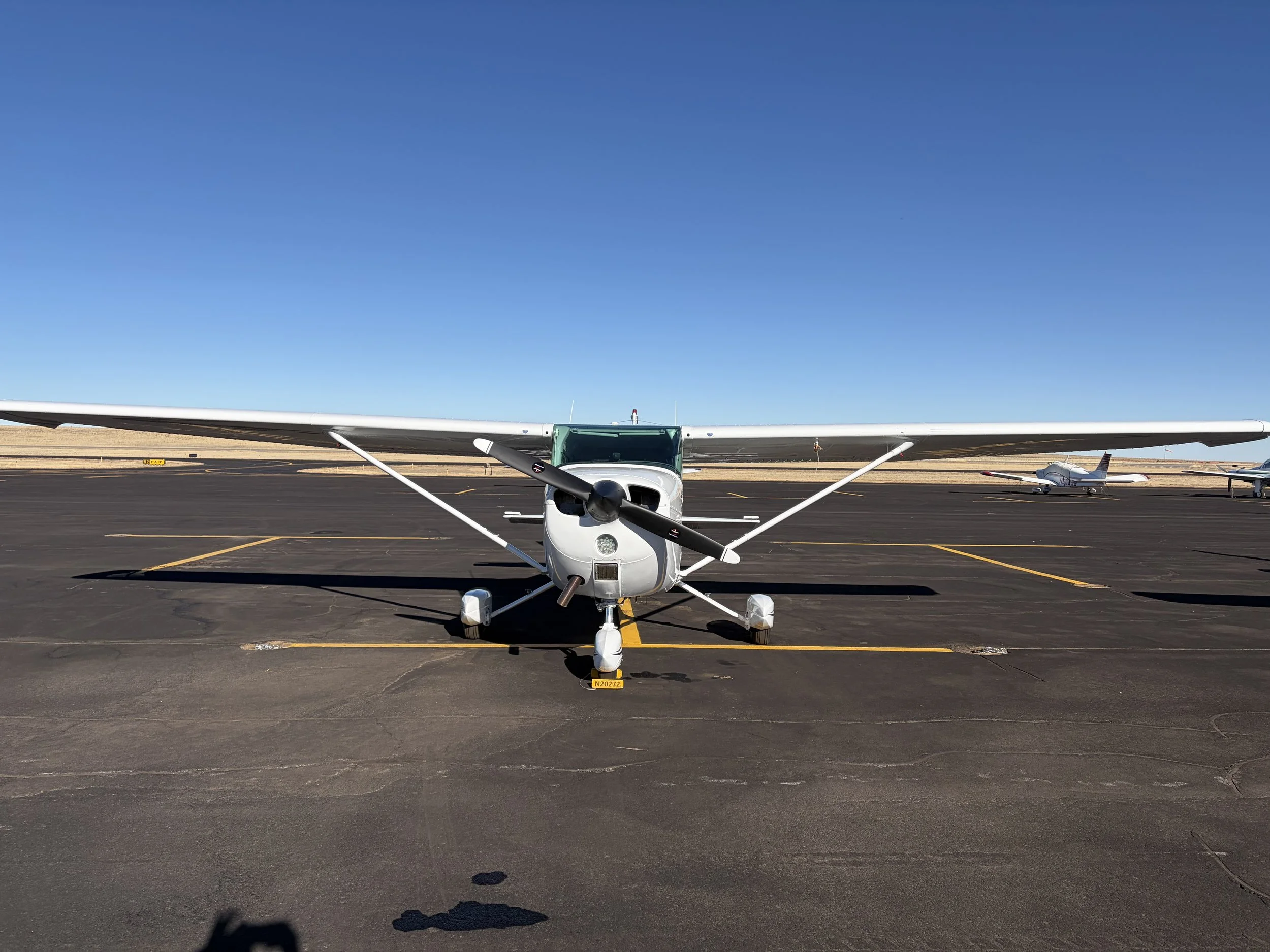 Front view of a small white airplane parked on a tarmac, with other airplanes in the background and a clear blue sky.