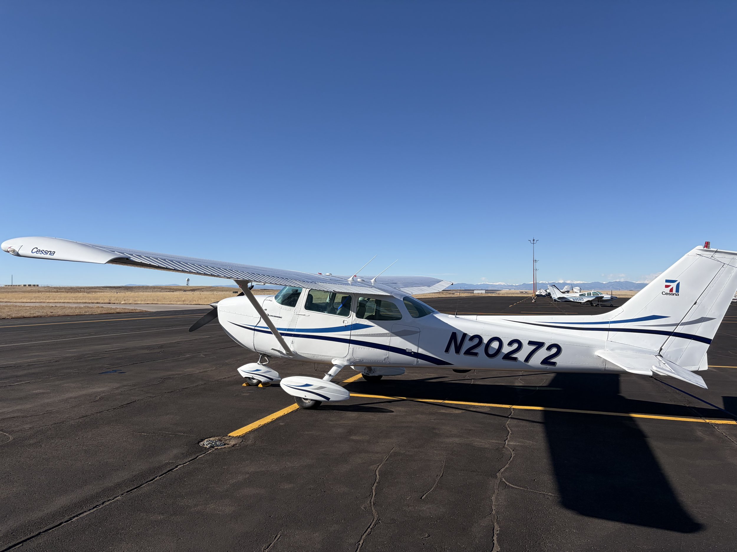 White Cessna airplane with blue and black stripes parked on airport tarmac under clear blue sky.