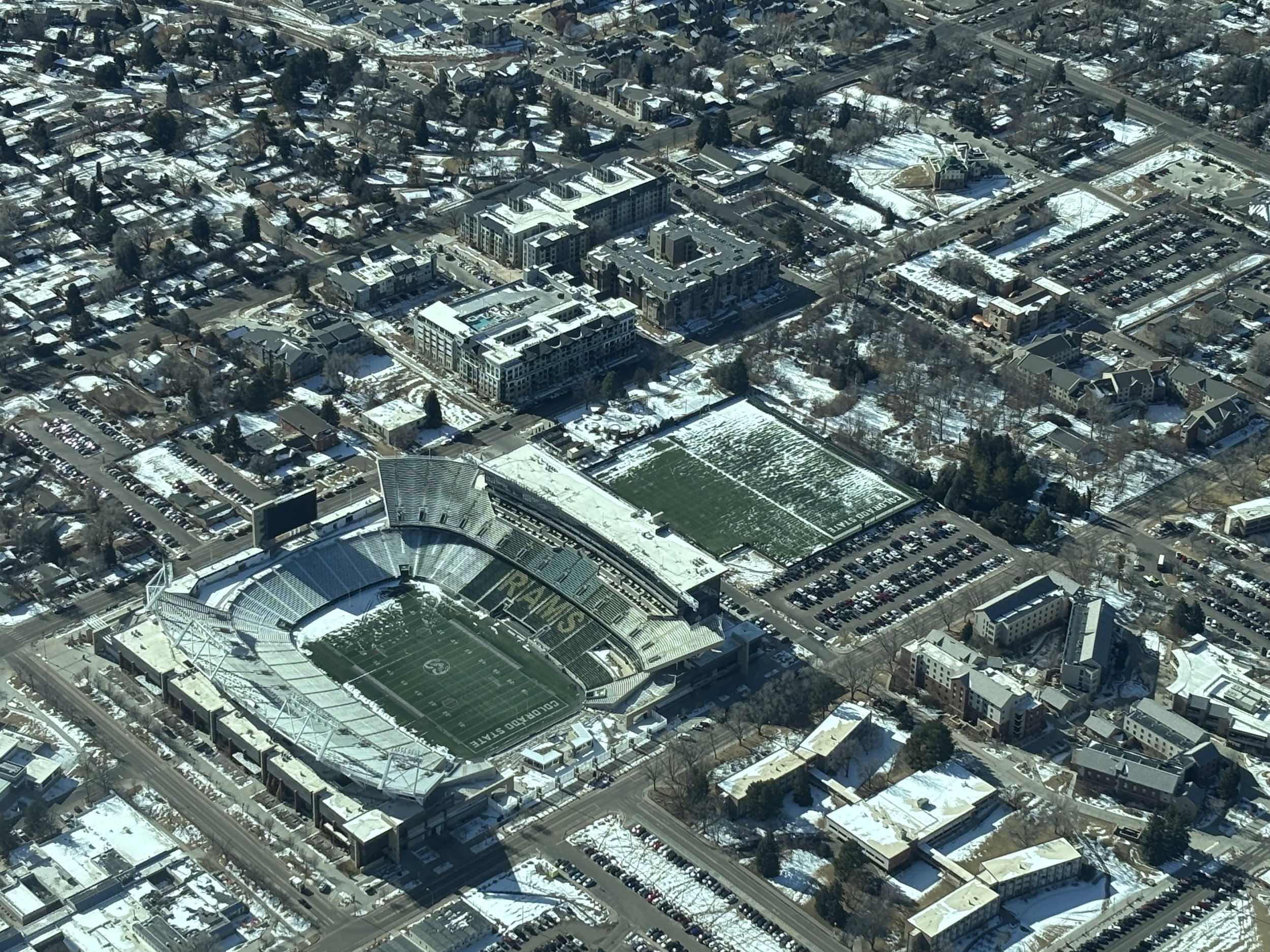 Aerial view of a football stadium with snow on the field and surrounding buildings, parking lots, and residential neighborhoods.