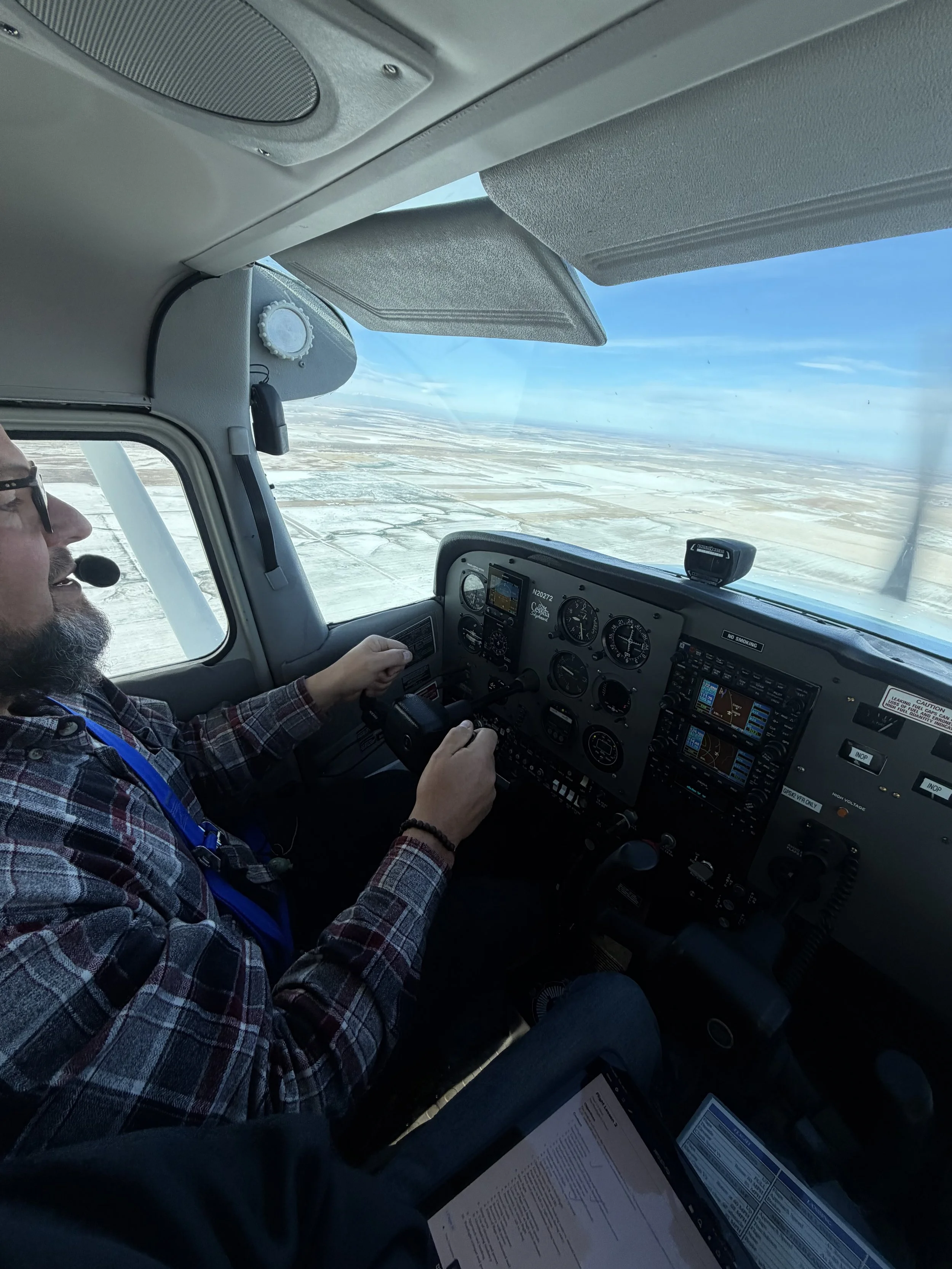 A pilot flying an airplane over snowy terrain with a clear blue sky, viewed from the cockpit.
