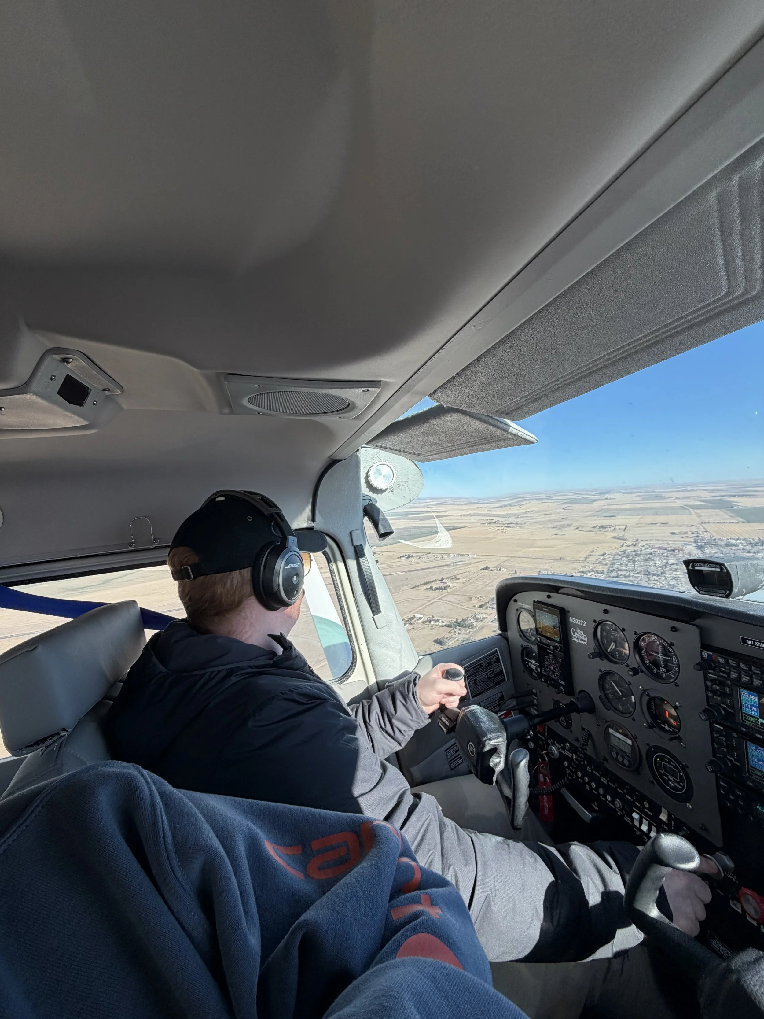 Pilot with headphones flying a small airplane over a flat rural landscape.