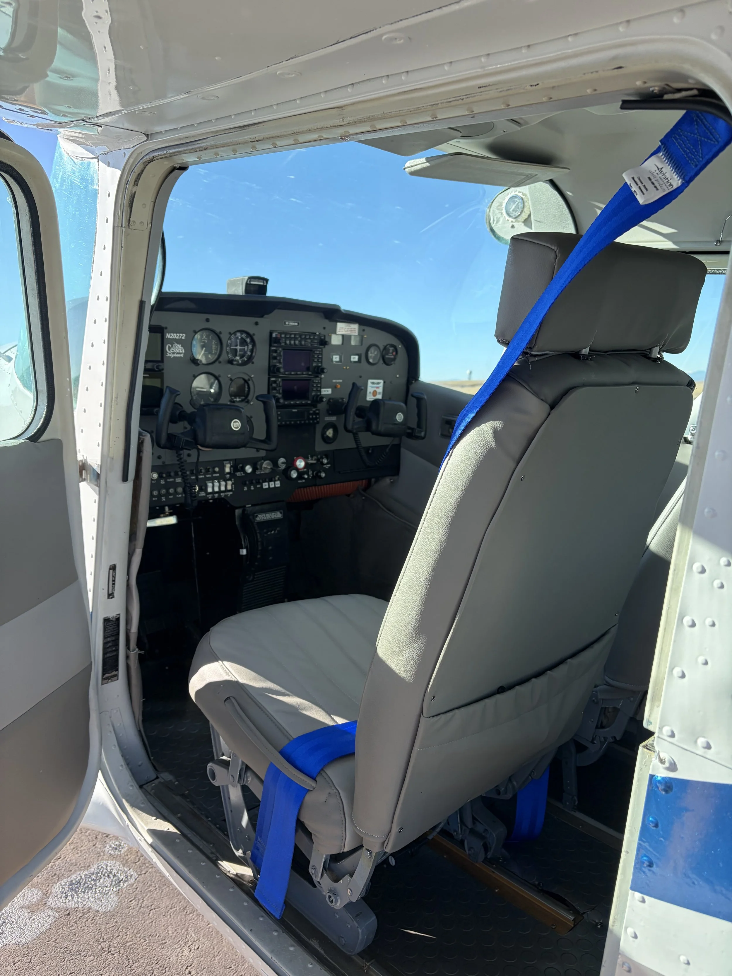 Interior view of a small aircraft cockpit with two front seats, flight instruments on the dashboard, and a large window showing a clear blue sky.