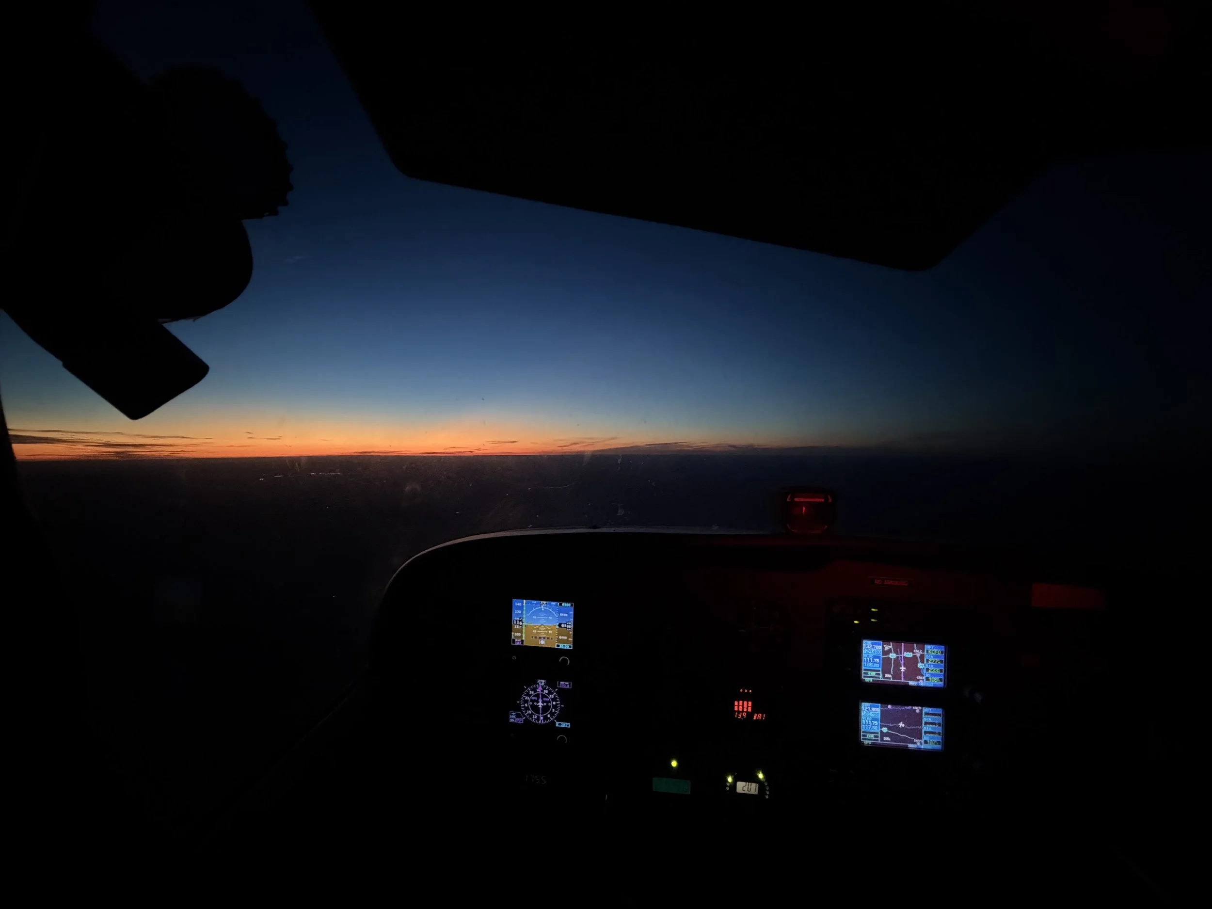 View from inside an airplane cockpit at twilight, showing the horizon with a colorful sunset sky and aircraft instruments illuminated.