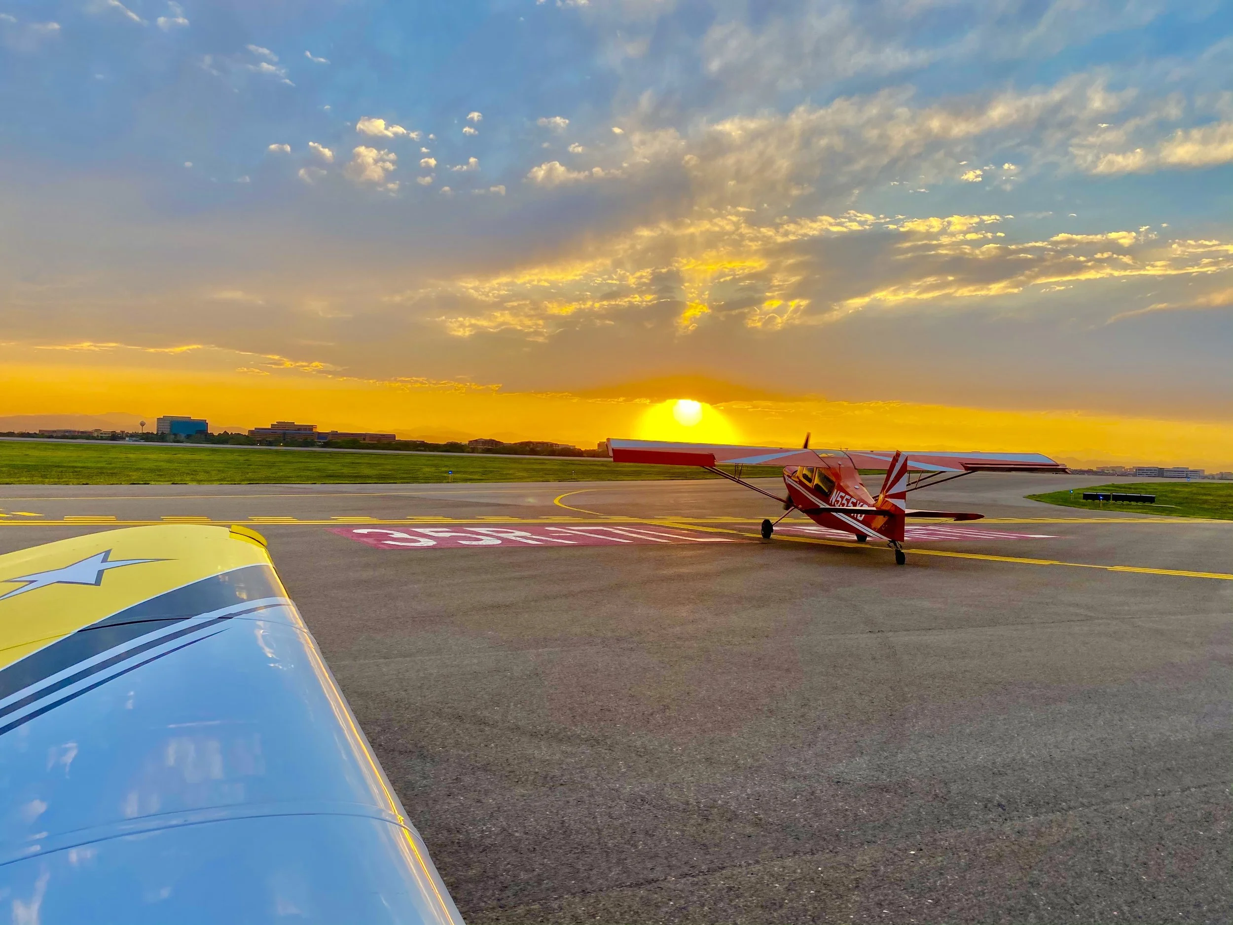 Small red and white aircraft on runway during sunset with colorful sky and clouds in background.