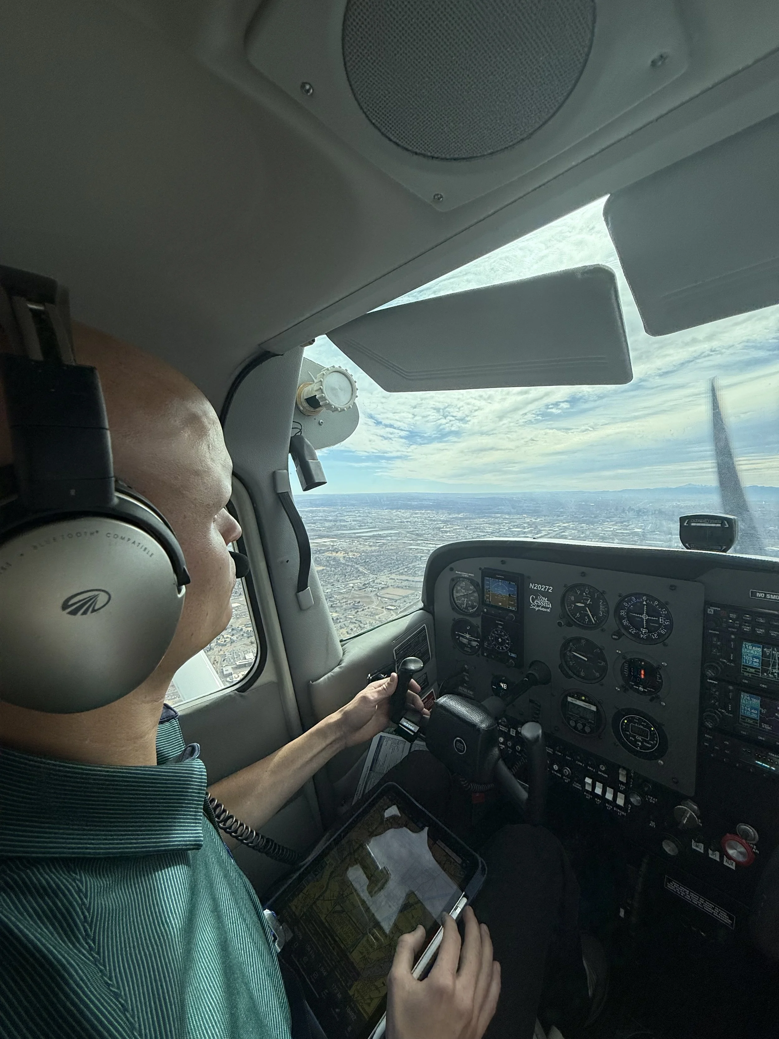 Pilot flying an aircraft, wearing a headset, holding a tablet, and controlling the airplane from the cockpit, with a view of the sky and landscape outside.