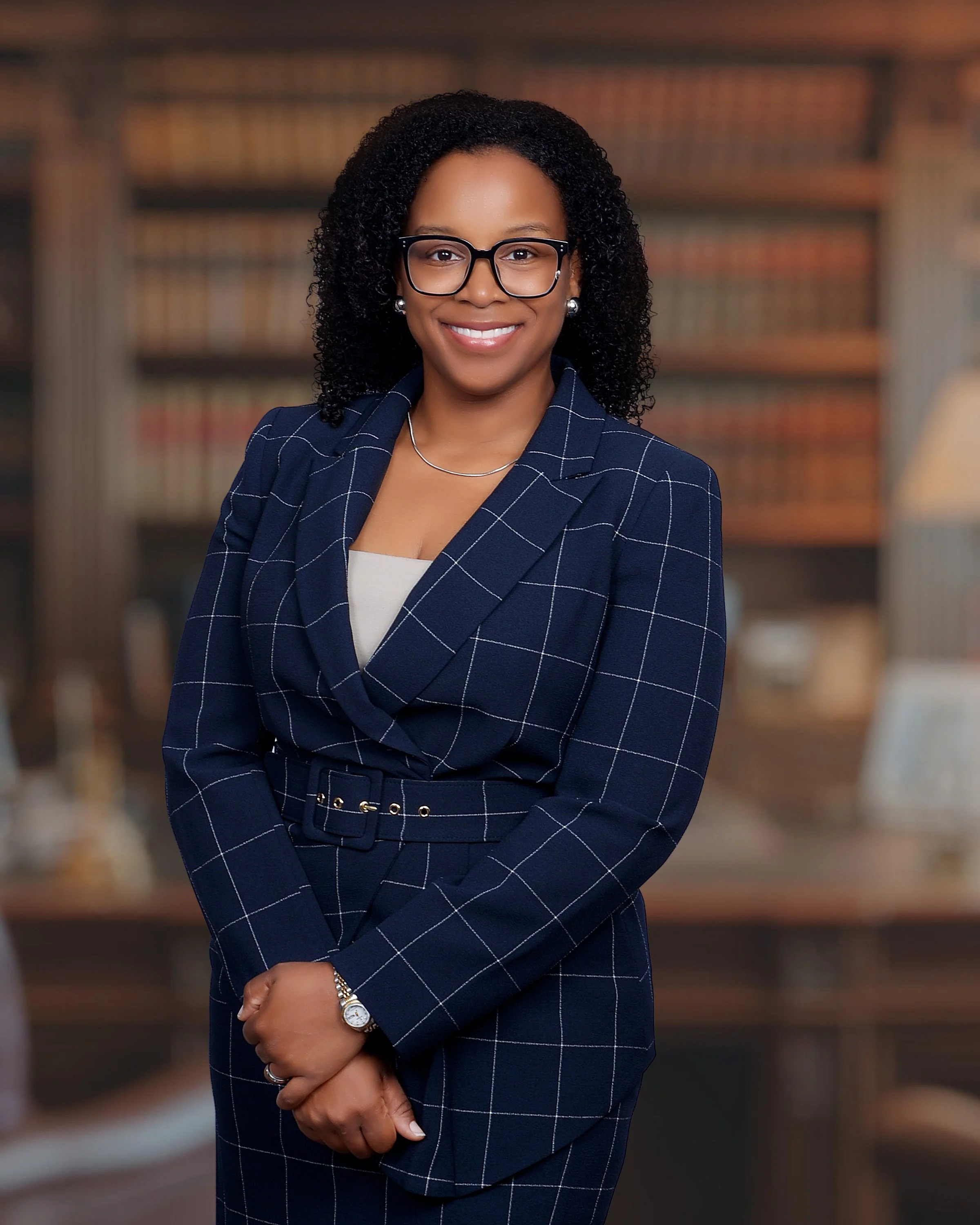 A professionally dressed woman smiling, standing in a library with bookshelves in the background.
