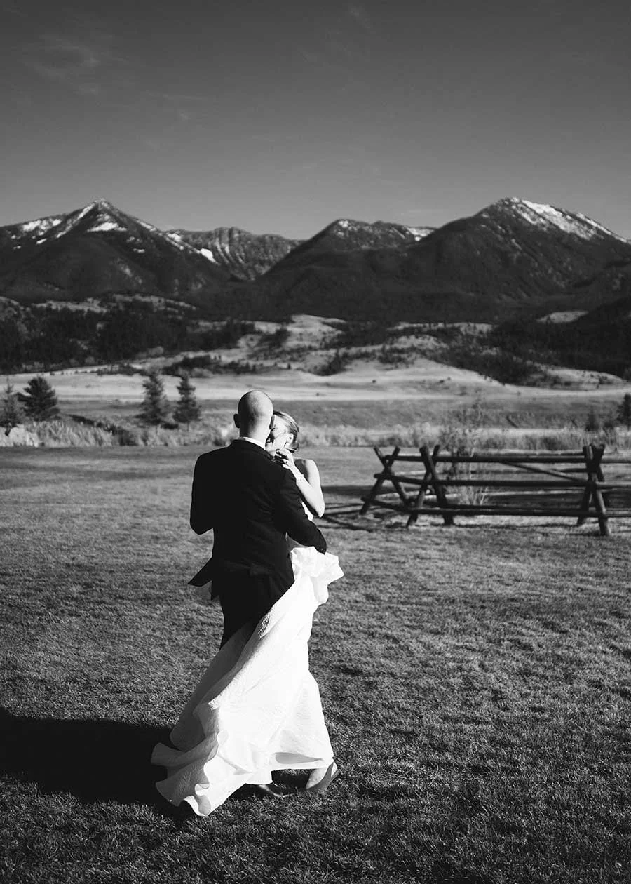 A black and white photo of a bride and groom dancing outdoors in a Montana mountain landscape, with snow-capped peaks in the background.