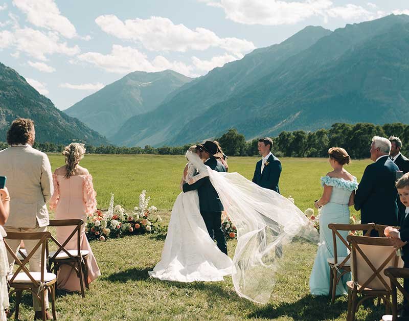 A Montana wedding ceremony outdoors with mountains in the background. A bride and groom are kissing in the center, surrounded by guests and floral arrangements.