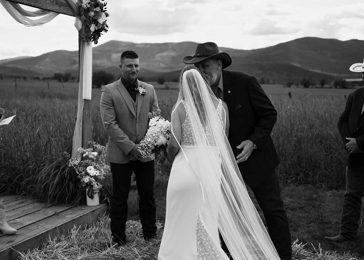 A wedding ceremony taking place outdoors on a grassy field, with mountains in the background. A bride and an older man wearing a cowboy hat are kissing, while a groom, dressed in a suit, stands nearby holding a bouquet of flowers.