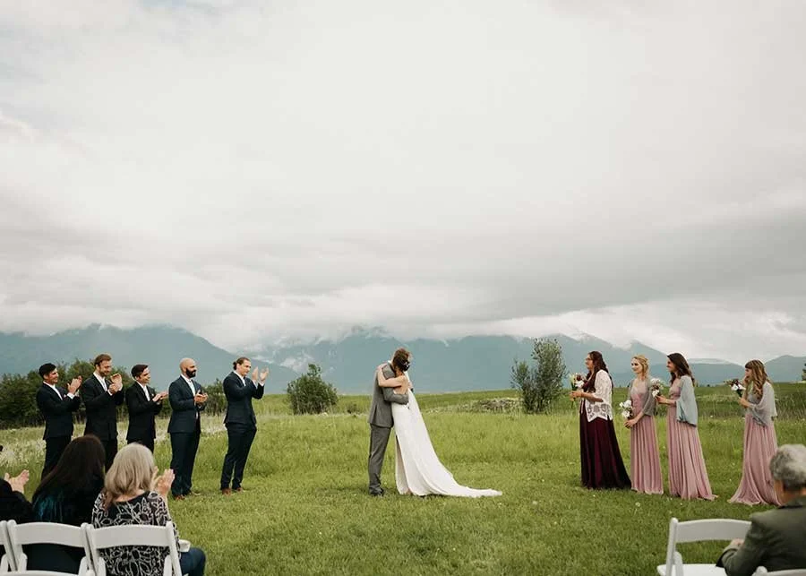 A couple kisses during an outdoor wedding ceremony on a grassy field with Montana mountains in the background, attended by friends and family.
