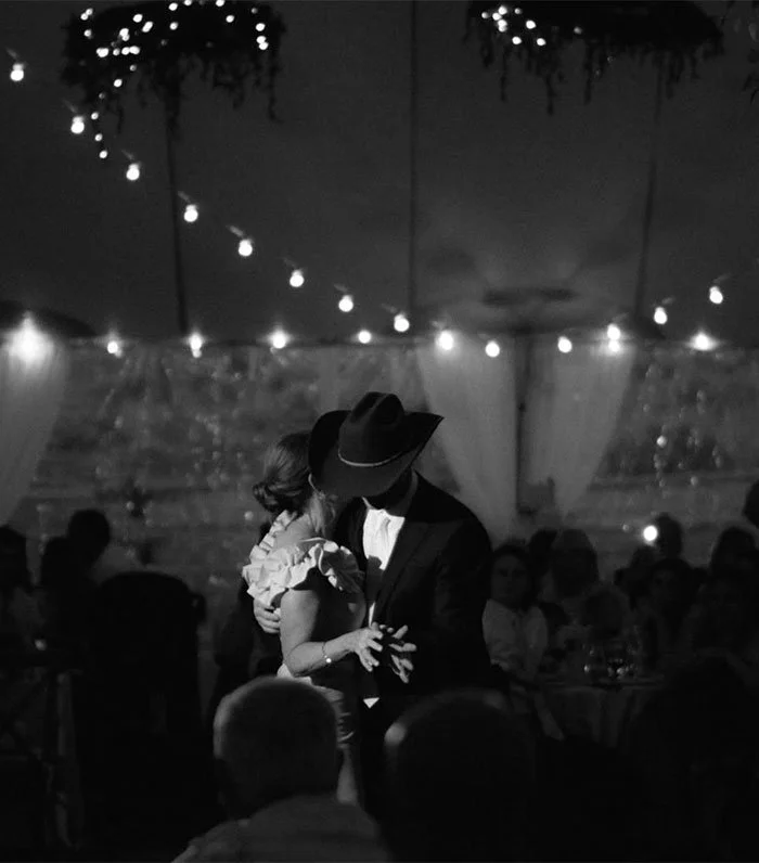 A couple dancing closely at a dimly lit event, with string lights overhead and decorated walls in the background.
