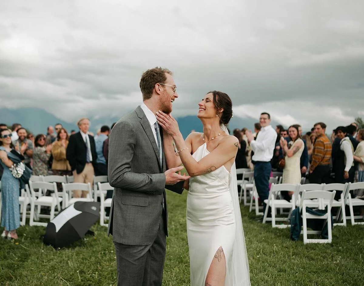 A bride and groom share a romantic moment during their outdoor wedding ceremony, smiling at each other as the bride touches the groom's face. Guests are seated behind them on chairs in a grassy outdoor setting with Montana sky overhead.