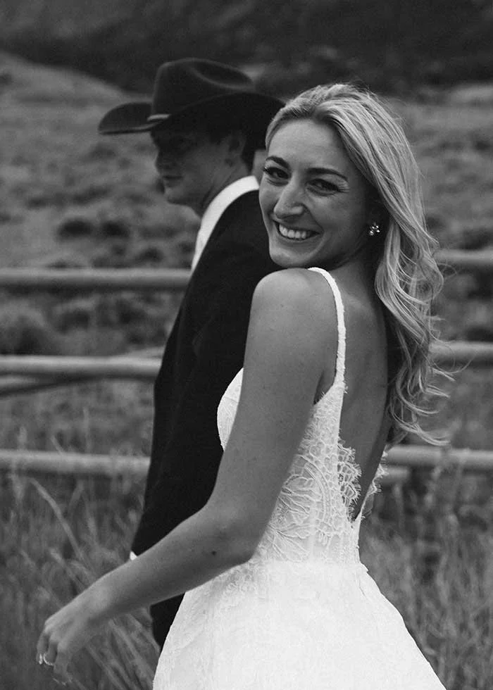 A smiling bride in a wedding dress outdoors, with her groom wearing a cowboy hat.